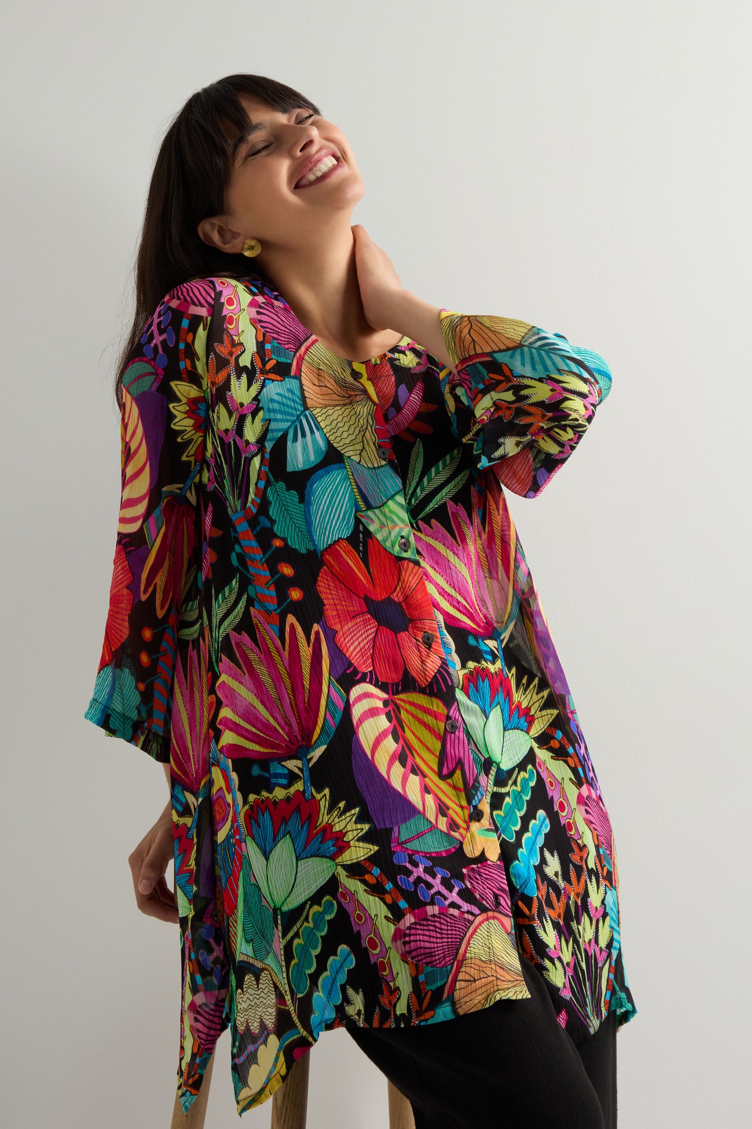 A woman smiles, tilts her head back, and wears the Fantasy Forest Georgette Shirt with its vibrant rainforest print, standing against a plain background for a chic jungle-luxe look.