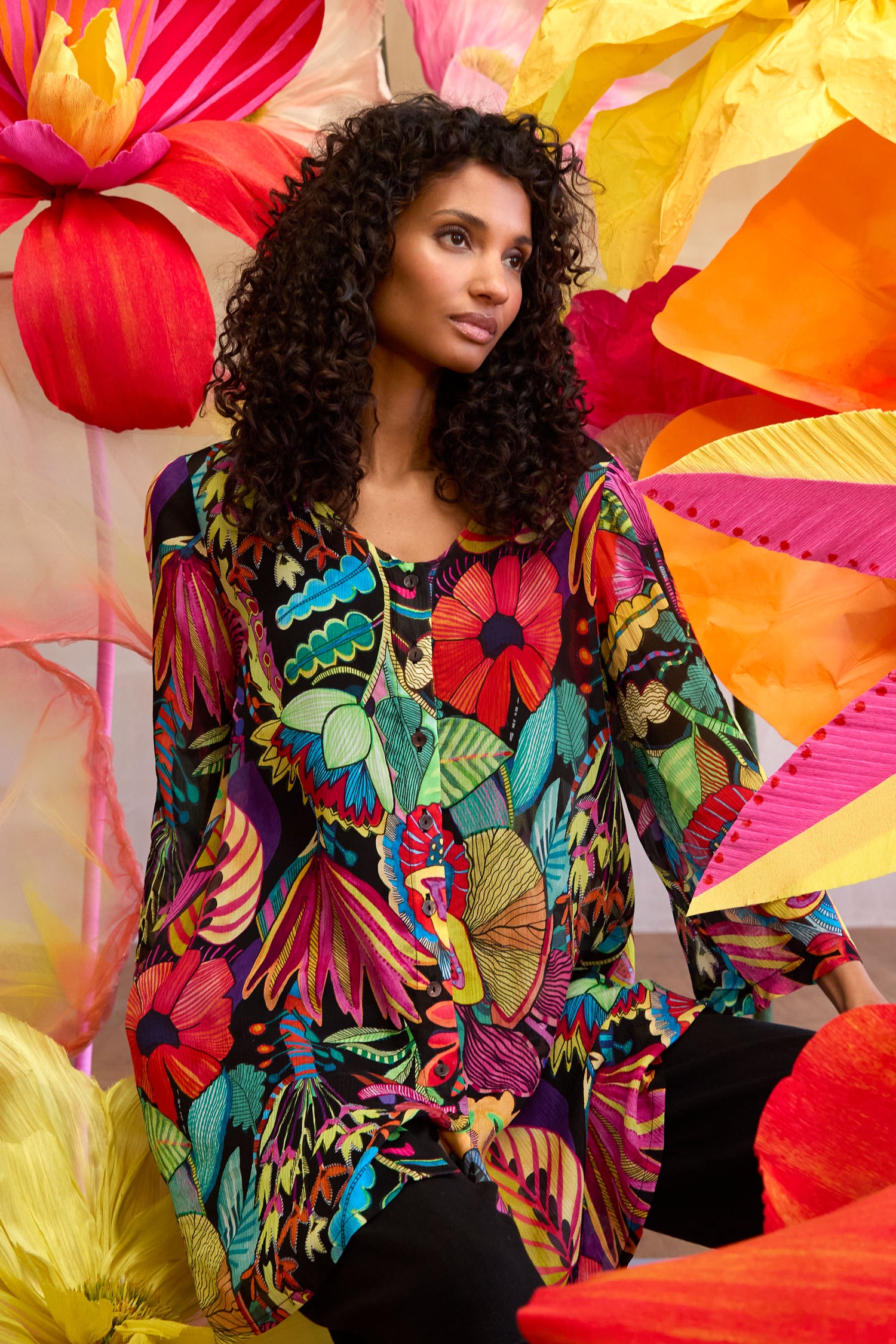 A woman with curly hair sits among oversized colorful paper flowers, wearing the Fantasy Forest Georgette Shirt featuring vibrant florals and lush leaves in a rainforest-inspired print.