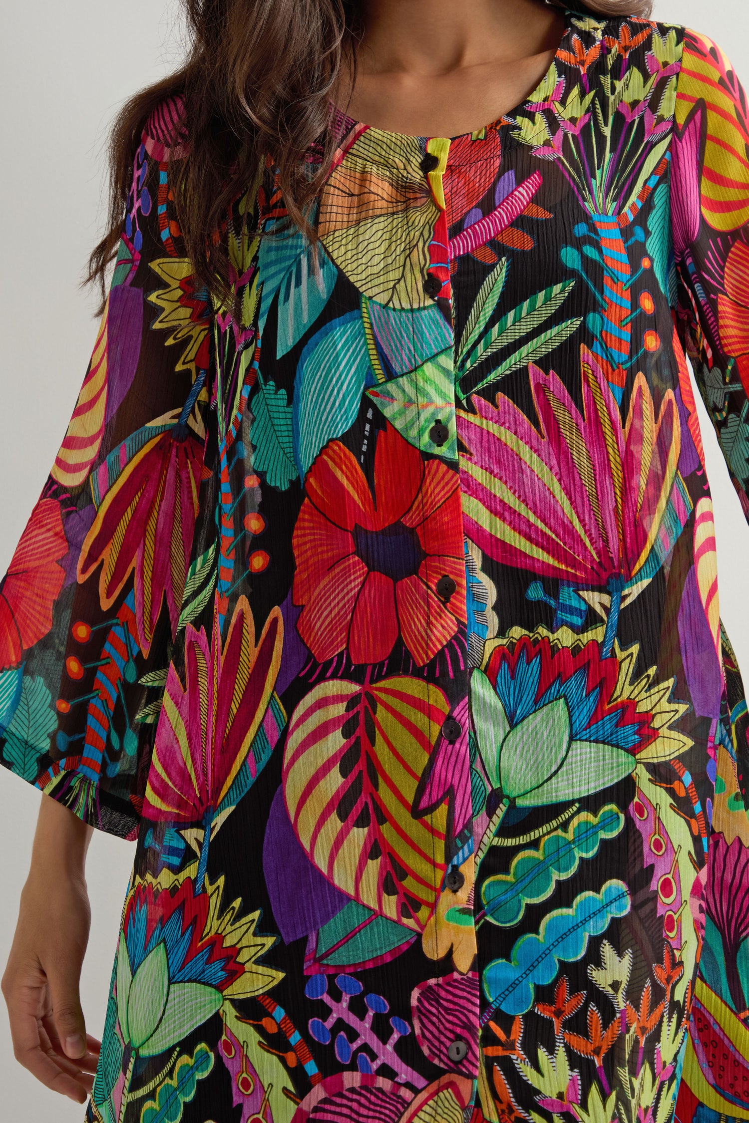 A person wearing the Fantasy Forest Georgette Shirt, featuring a jungle-luxe, bold tropical floral print in vivid red, pink, blue, green, and yellow on black.