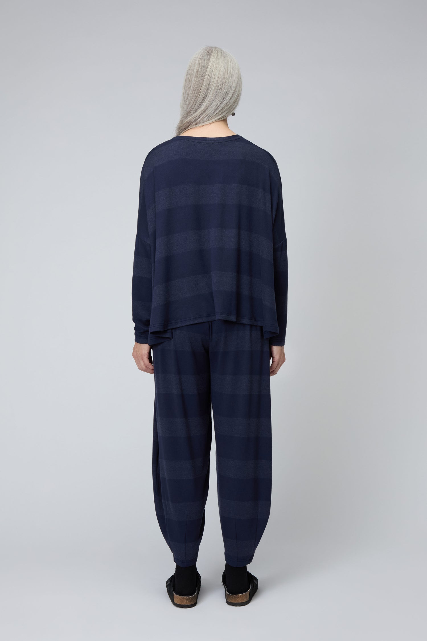 A person with long gray hair models the Soft Viscose Jersey Stripe Boxy Top—loose-fitting and navy—paired with matching pants, barefoot and facing away from the camera against a plain light background. Made in England.