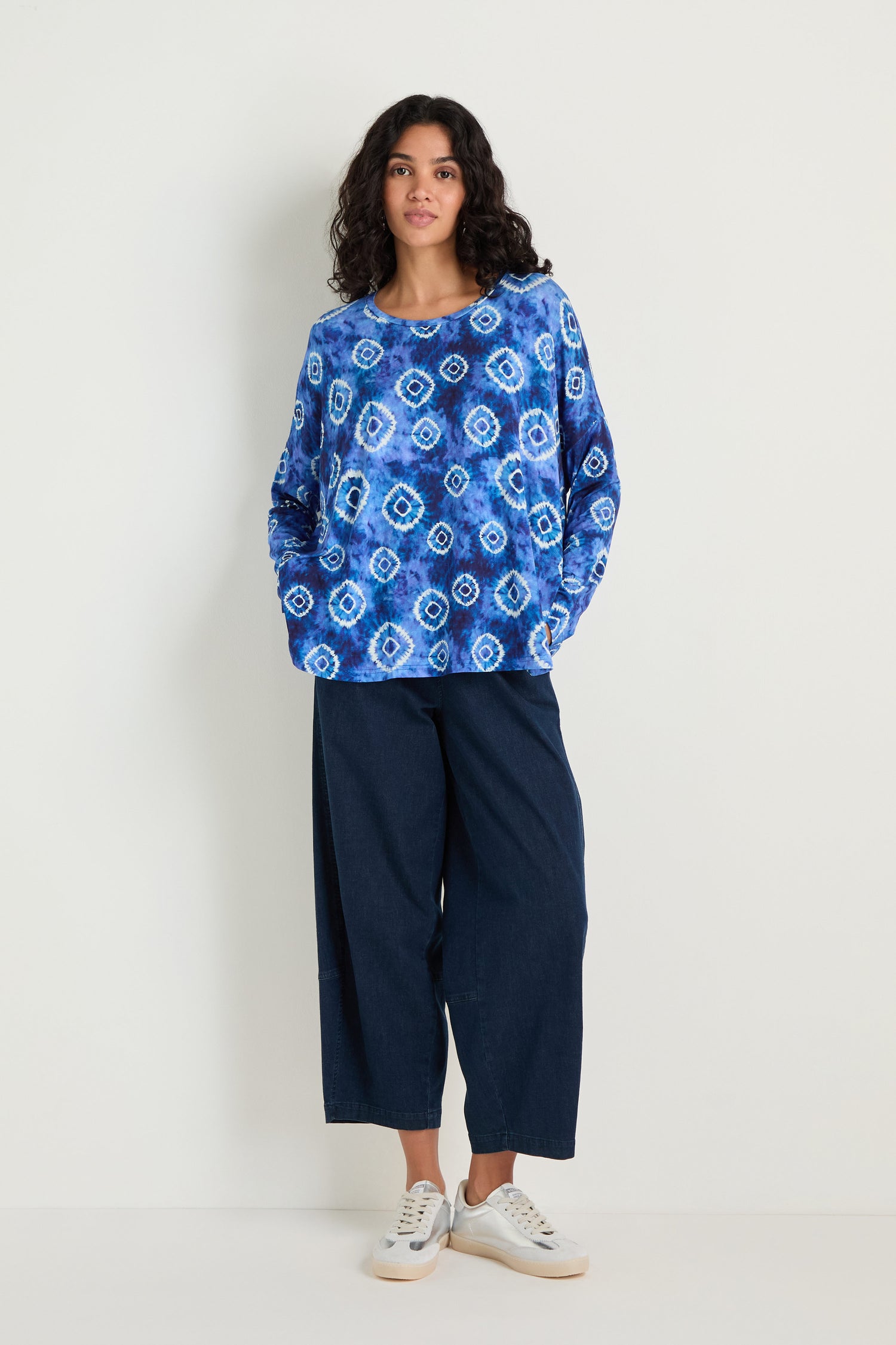 A person stands against a plain white wall wearing the Tie Dye Circles Jersey Boxy Top, wide-leg dark blue pants, and white sneakers.