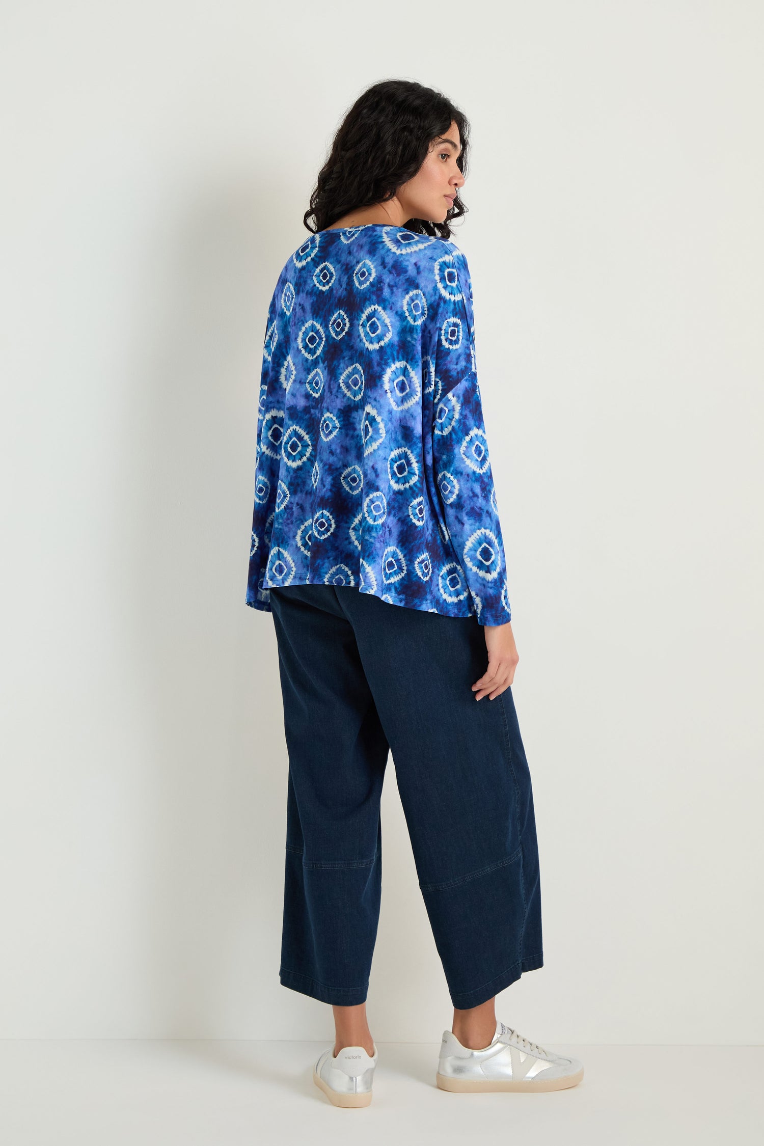 A person faces away, wearing the Tie Dye Circles Jersey Boxy Top with wide-leg dark jeans and white sneakers against a plain light background.