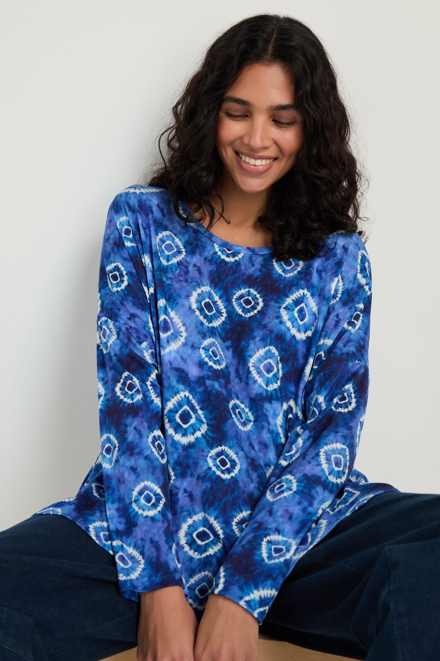 A smiling woman with curly hair wears the Tie Dye Circles Jersey Boxy Top, featuring blue geometric circle motifs in soft viscose jersey, set against a plain light background.