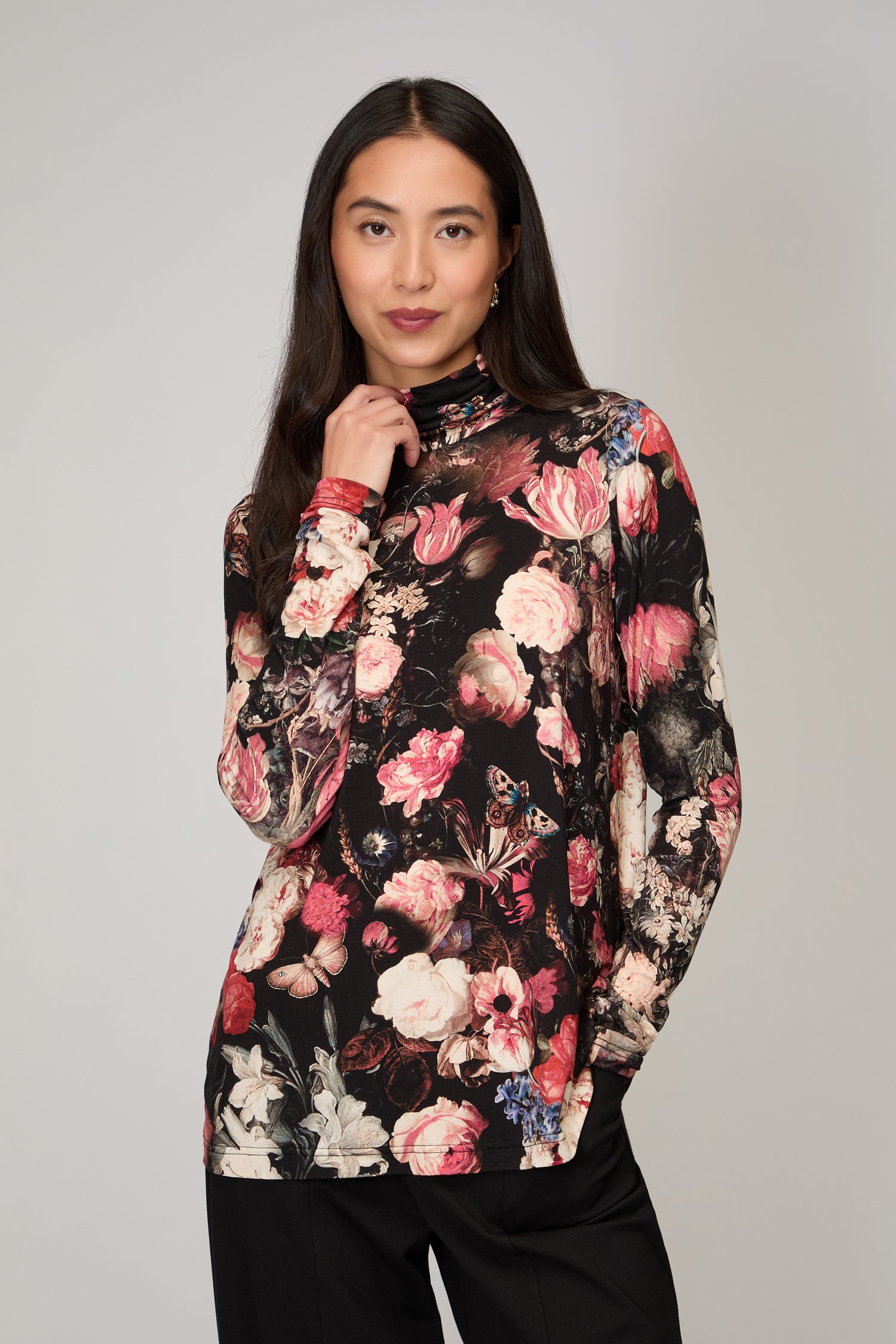 A woman with long dark hair wears a black Renaissance Floral Jersey Roll Neck Top and black pants, standing against a plain grey background.