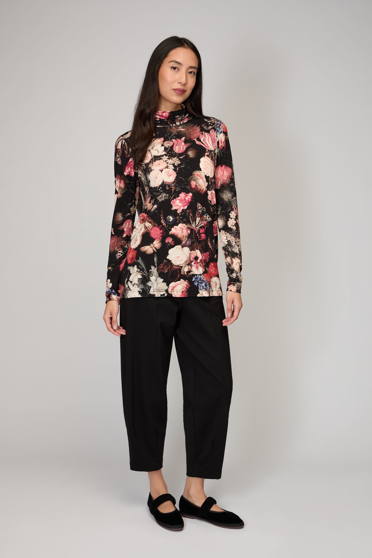A woman stands against a plain background wearing the Renaissance Floral Jersey Roll Neck Top in printed viscose jersey, styled as a chic layer with black ankle-length pants and black shoes.