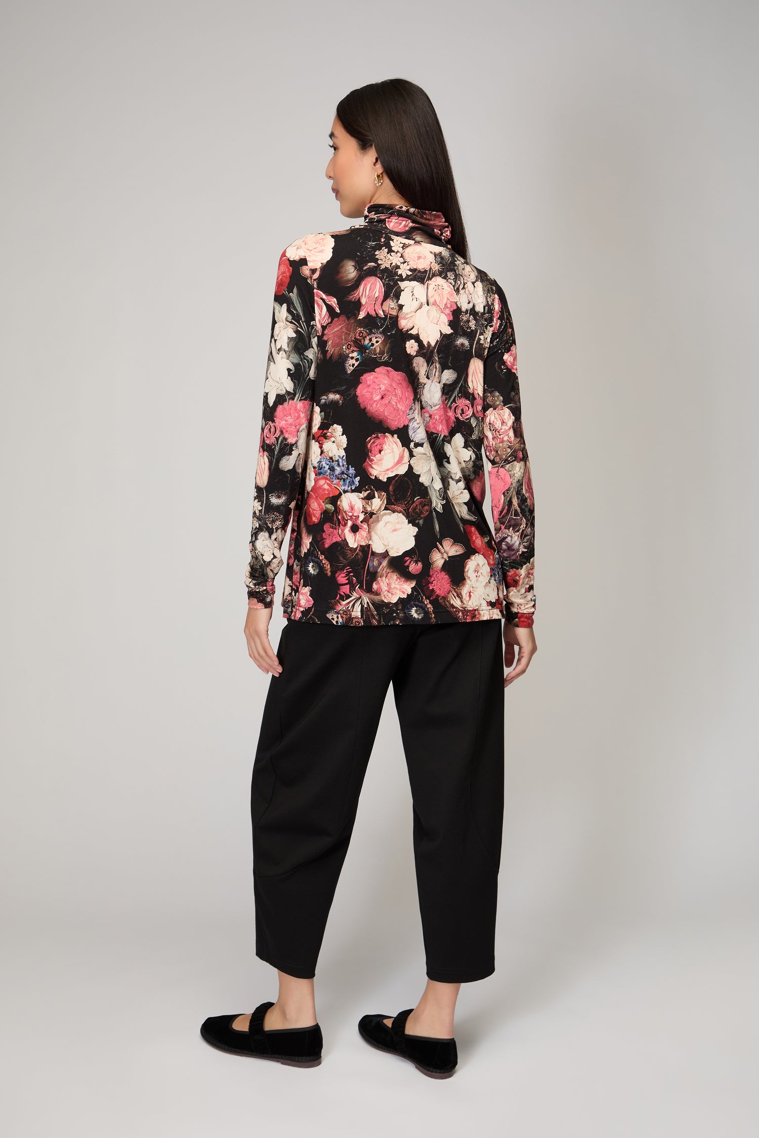 A woman faces away, wearing the Renaissance Floral Jersey Roll Neck Top with black pants and shoes against a plain light background—the ideal layering piece for any wardrobe.