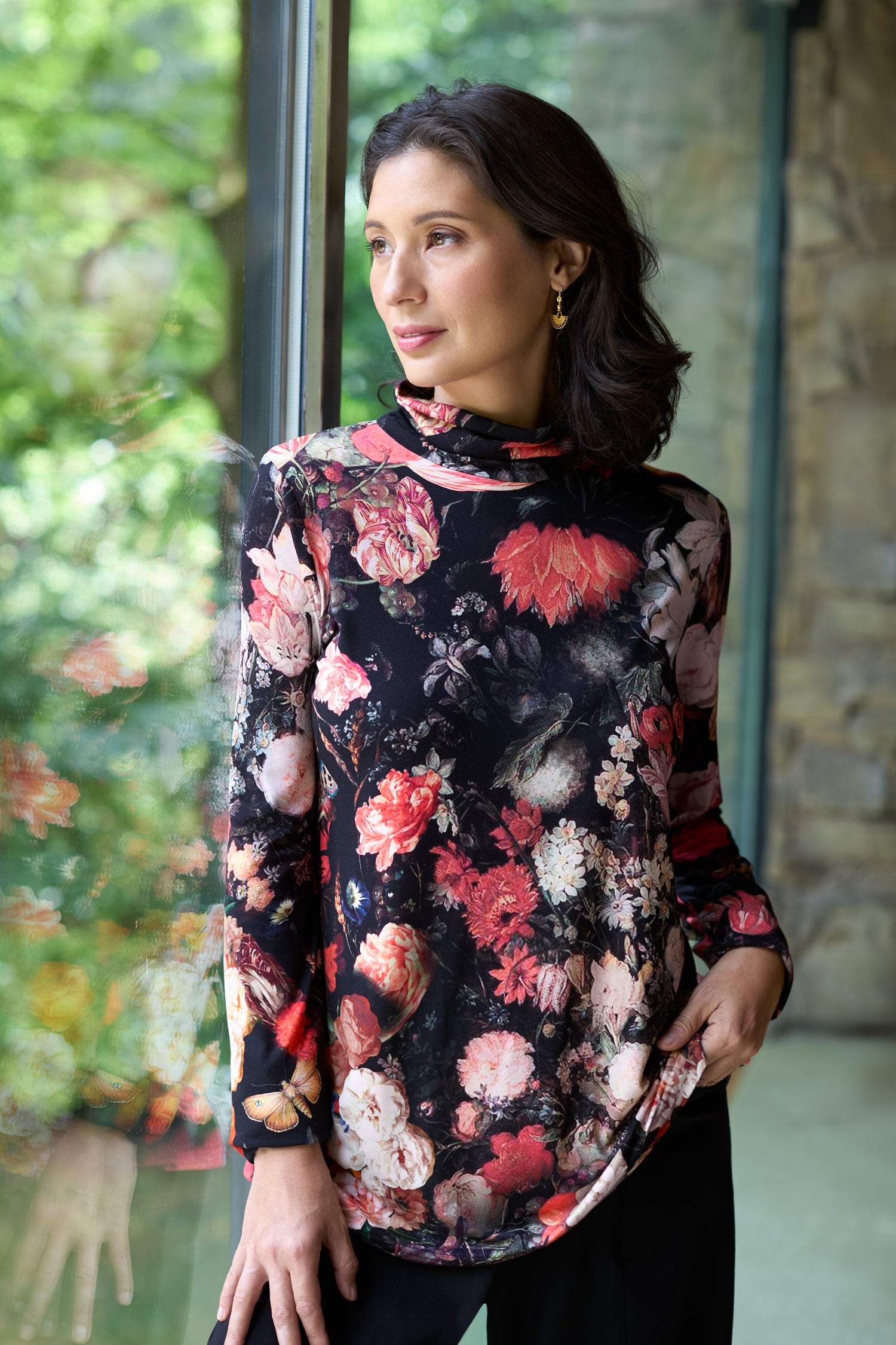A woman stands by a window in the Renaissance Floral Jersey Roll Neck Top, made in the UK, paired with black pants. She looks to the side with one hand in her pocket.