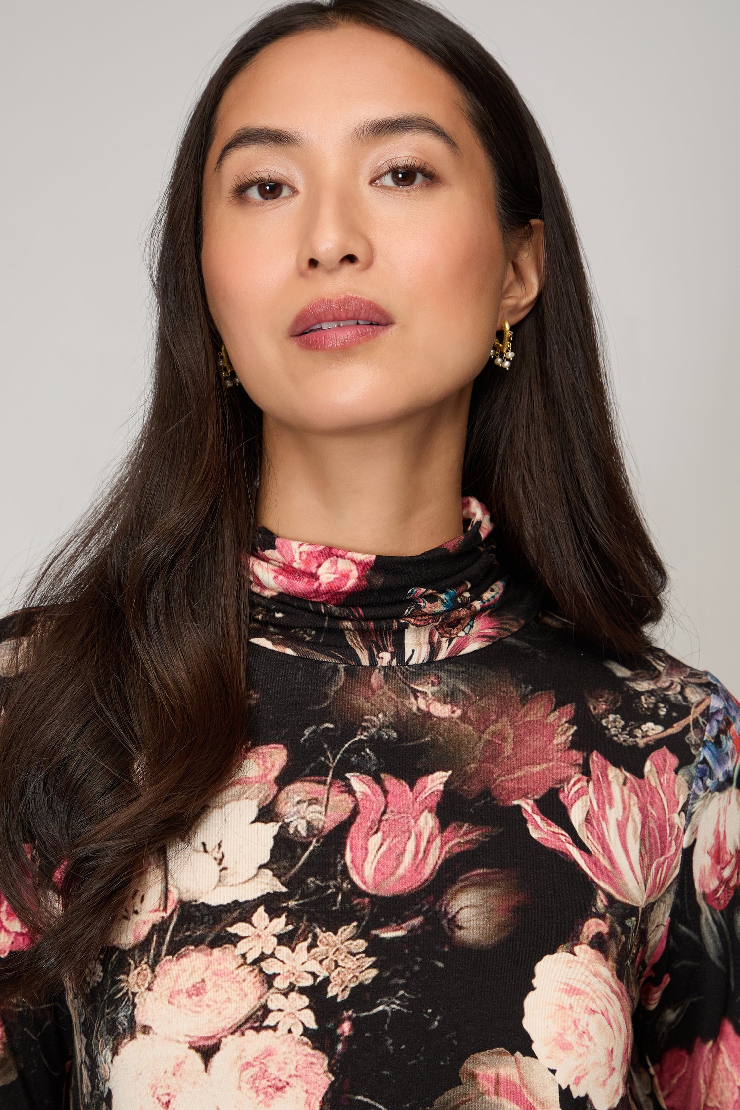 A woman with long dark hair wears the Renaissance Floral Jersey Roll Neck Top in printed viscose jersey and gold earrings, looking at the camera against a plain background—an easy layering piece for any season.