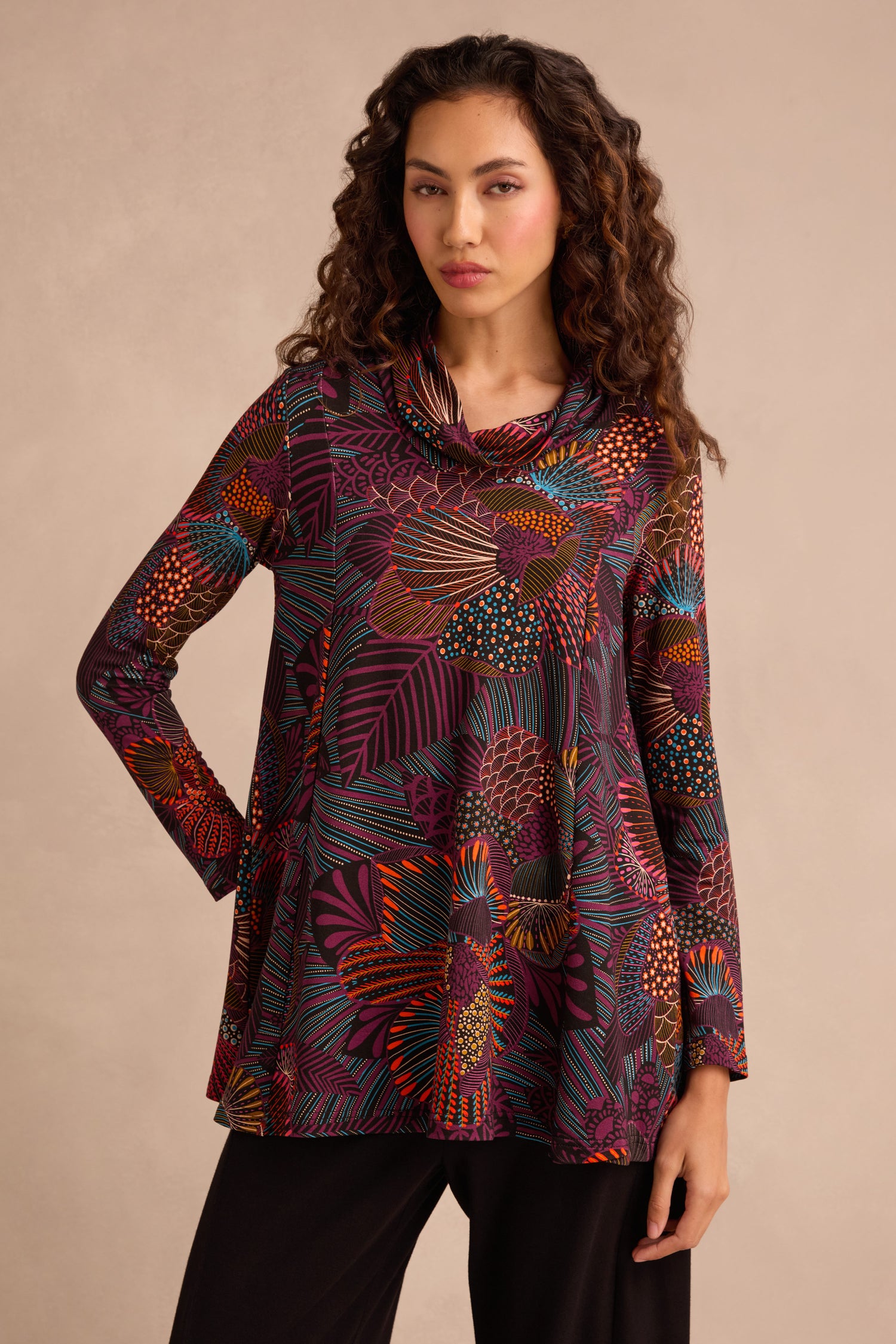 A woman is wearing the Abstract Linear Floral Jersey Tunic, featuring a long-sleeve, fit and flare silhouette with abstract floral and leaf patterns in red, orange, purple, and yellow shades. She has paired it with black pants and stands against a neutral background.