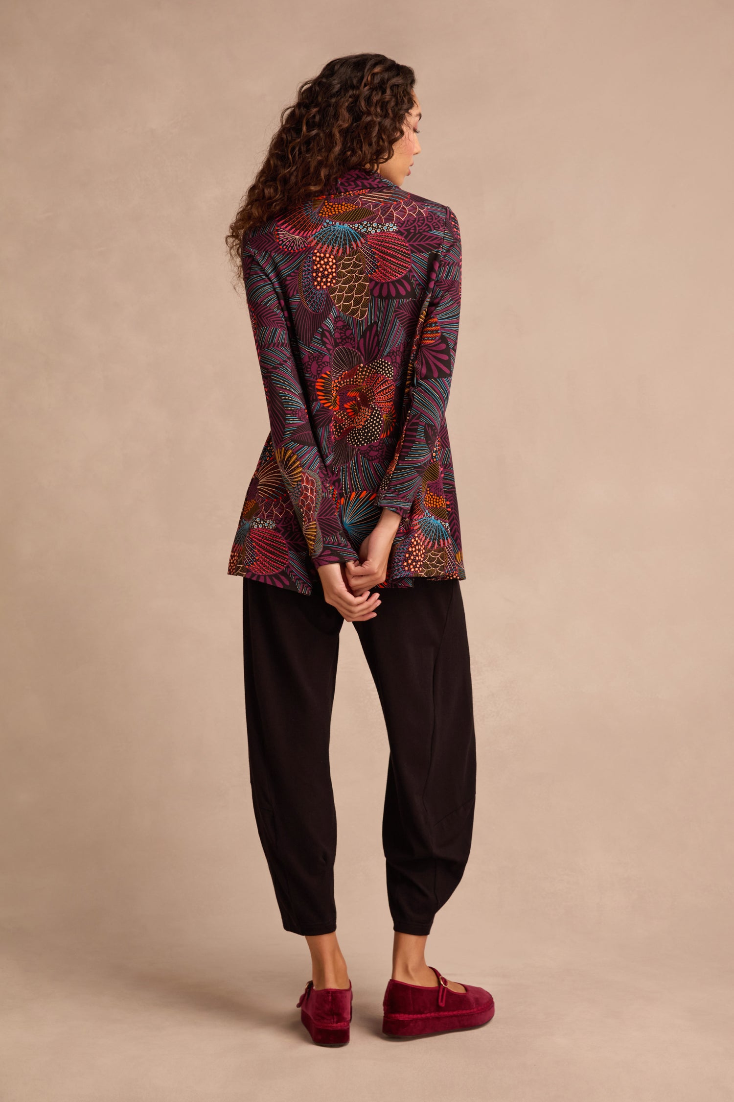 A person stands facing away, wearing a patterned blazer layered over the Abstract Linear Floral Jersey Tunic—with its cowl neck and fit-and-flare silhouette—paired with black pants and red slip-on shoes, hands clasped behind their back against a beige background.