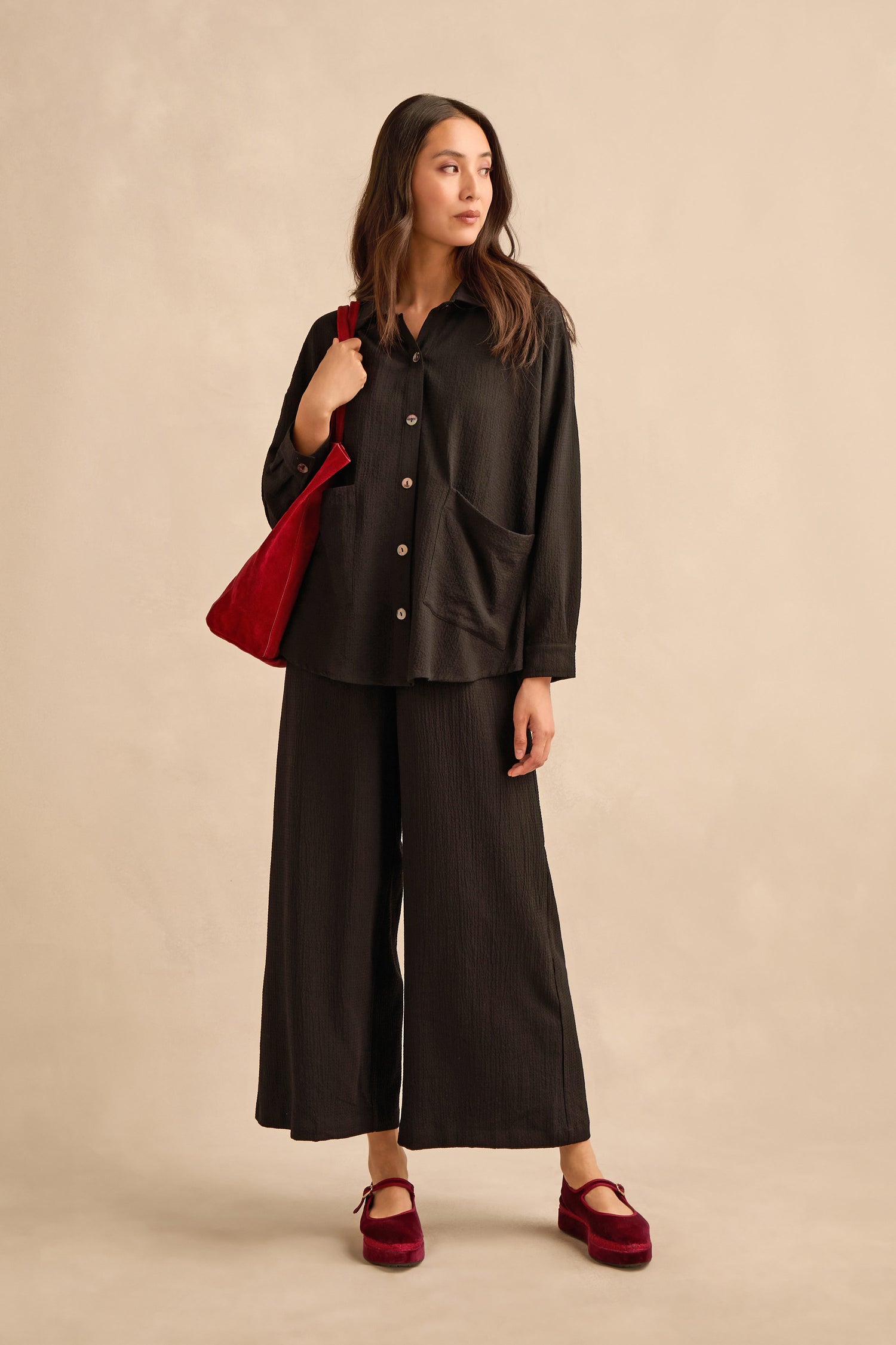 A woman stands facing left, wearing the Mini Crinkle Viscose Boxy Shirt in black paired with wide-leg pants, red velvet shoes, and a red shoulder bag, set against a neutral beige background.