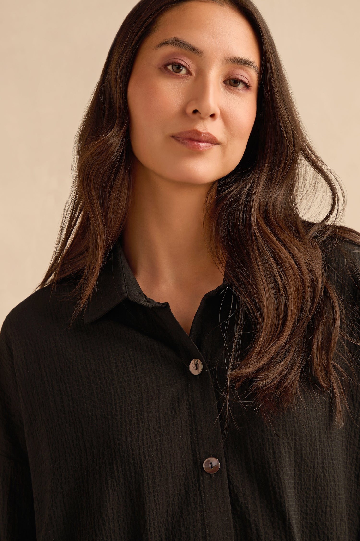 A woman with long brown hair wears the Mini Crinkle Viscose Boxy Shirt, standing against a neutral background and gazing slightly to the side with a calm expression.
