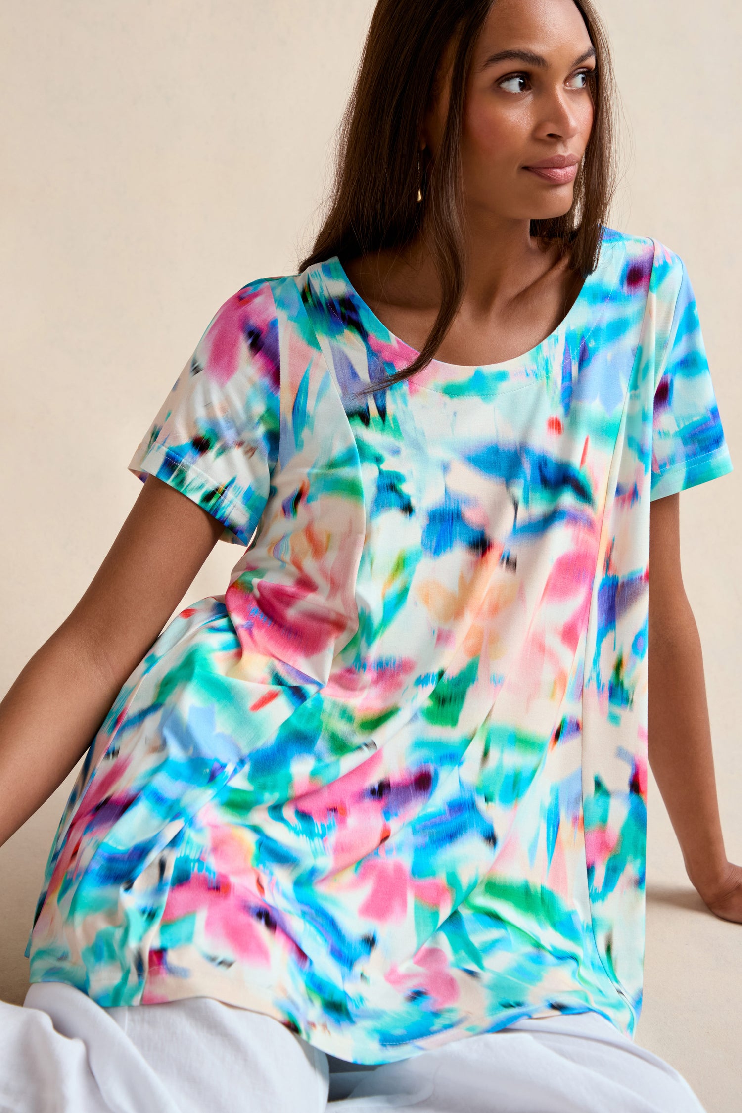 A woman sits wearing the Petal Haze Jersey Tunic, a relaxed fit, short-sleeve top featuring a colorful abstract watercolor pattern.