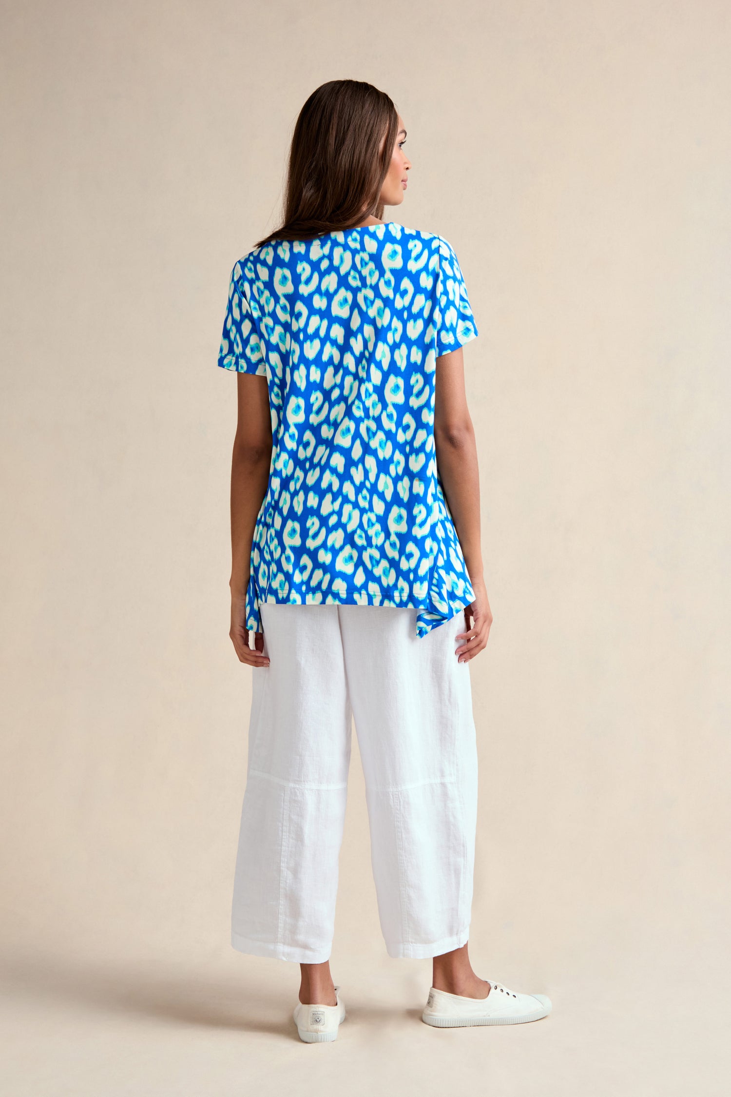 A woman stands facing away, wearing an Abstract Leopard Print Jersey Top with white wide-leg pants and white sneakers against a plain beige background.