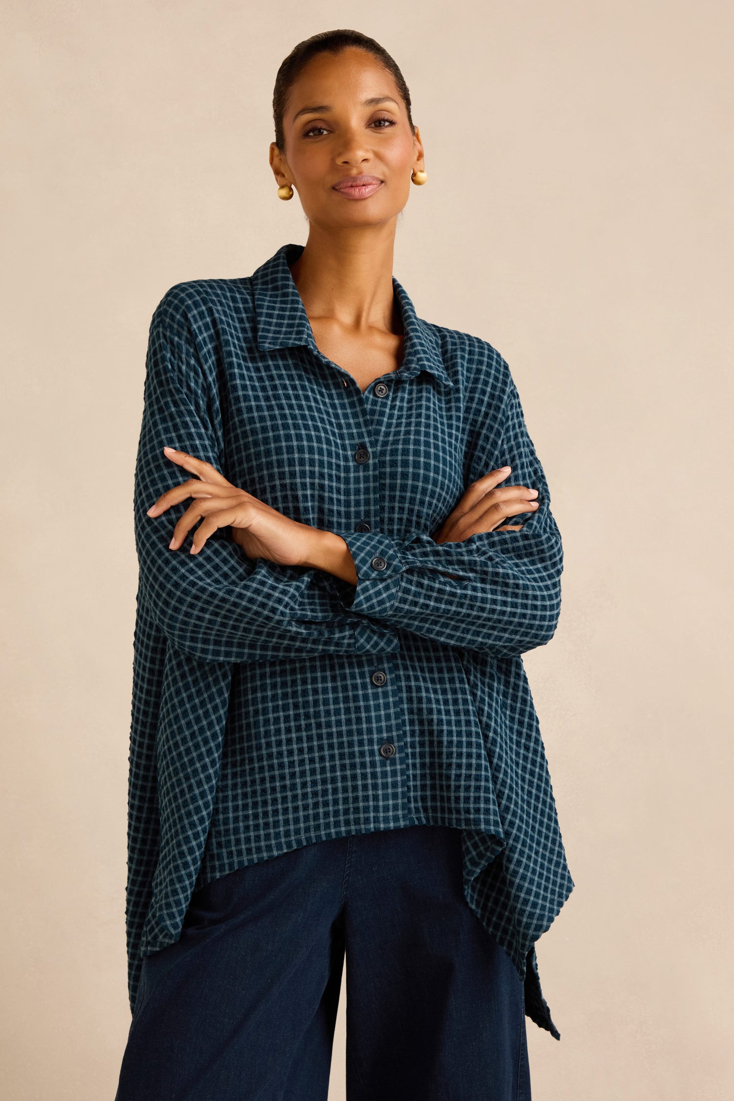 Model wears the Seersucker Squares Relaxed Shirt—featuring a blue checkered pattern and boxy silhouette—paired with dark wide-leg pants, standing with arms crossed and facing the camera against a neutral background.