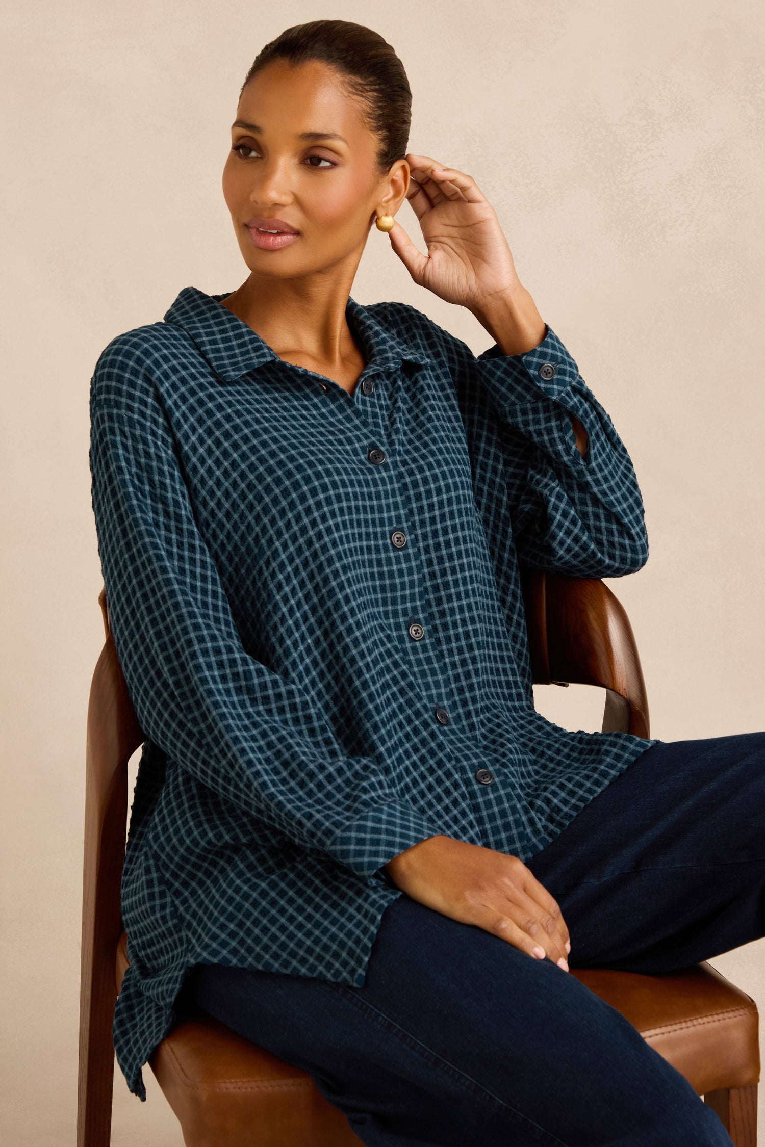 A woman sits on a chair wearing the Seersucker Squares Relaxed Shirt, which features a blue checkered pattern and a boxy silhouette, paired with dark pants. She touches her ear with one hand and gazes to the side.