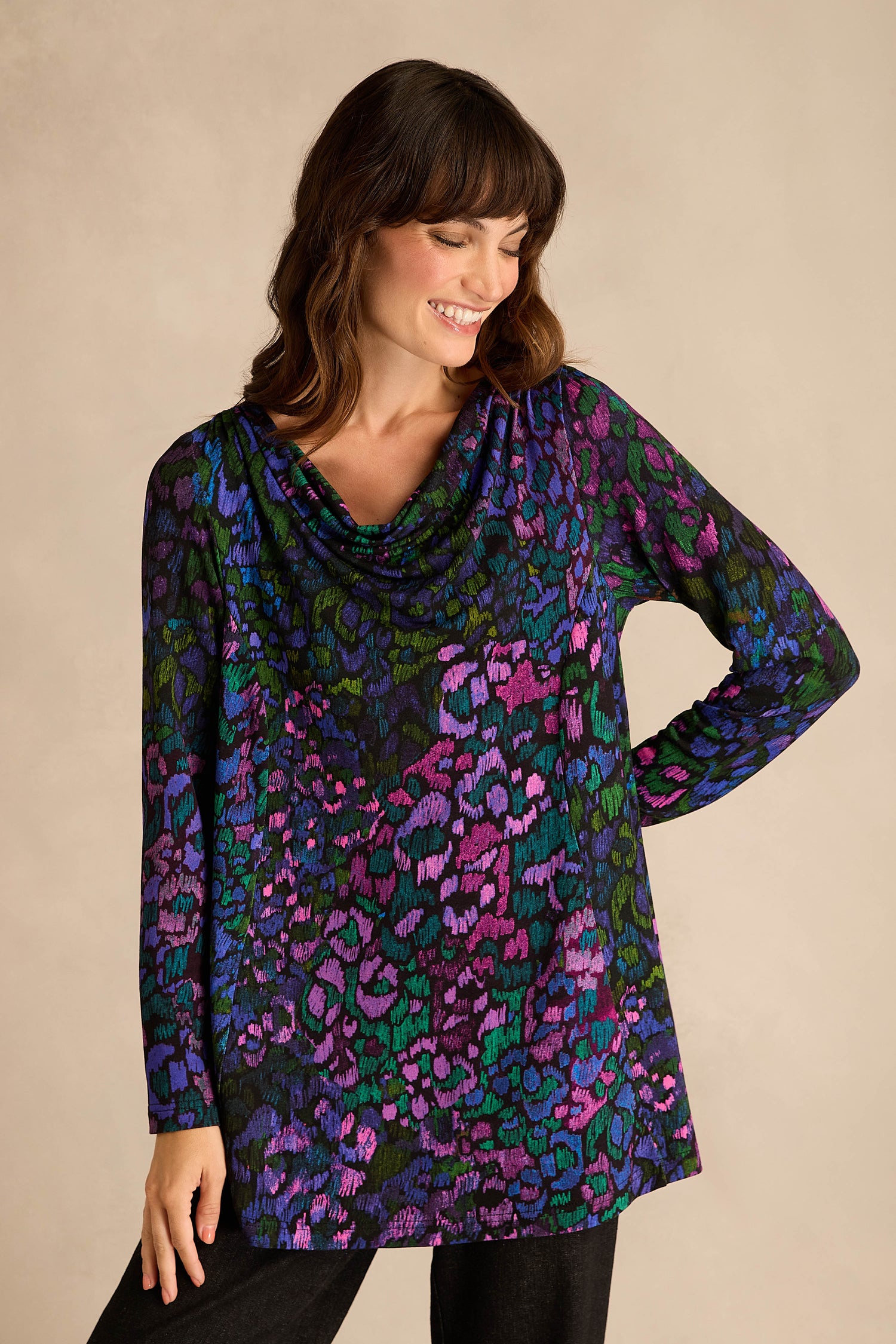 A woman stands against a plain background, wearing the Winter Garden Jersey Cowl Neck Top, which features long sleeves and a colorful abstract pattern in blue, green, purple, and black. With its flattering fit-and-flare silhouette and stylish cowl neck, she looks down with a slight smile.