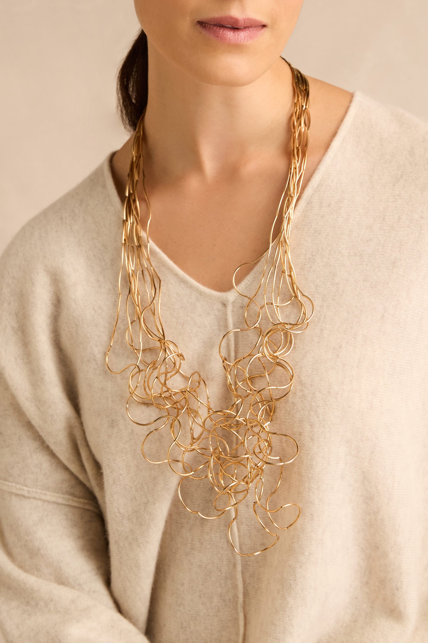 A woman shown from the shoulders up against a neutral background wears a cream sweater paired with the Gold Long Medley Necklace.