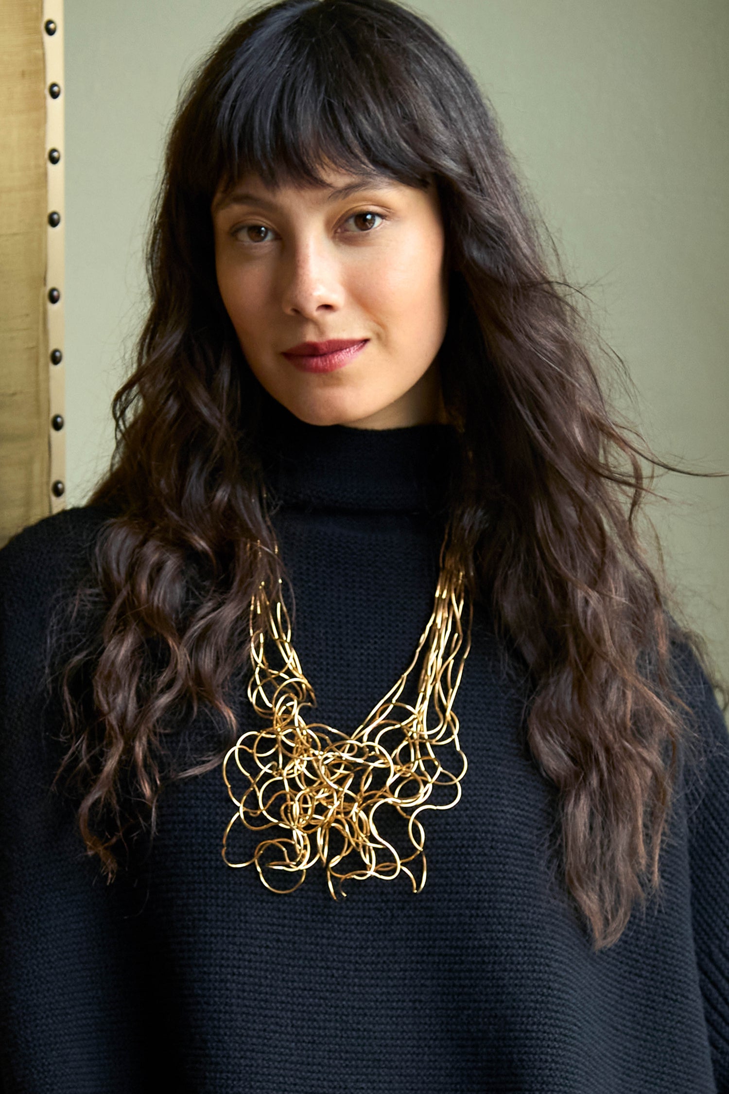 A person wearing a black turtleneck sweater and a Gold Long Medley Necklace featuring a cluster of interconnected wire loops.