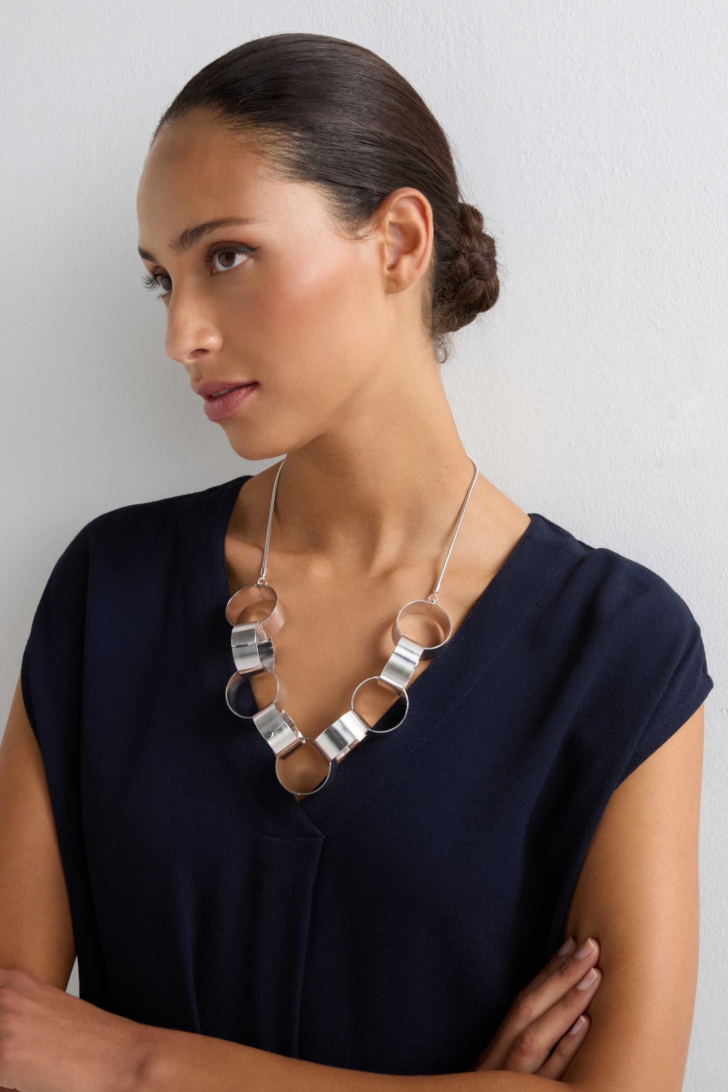 Woman with dark hair in a low bun wearing a navy V-neck top and the Affinity Hoop Necklace, which features a sculptural chunky design with a brushed silver finish, standing against a white background.