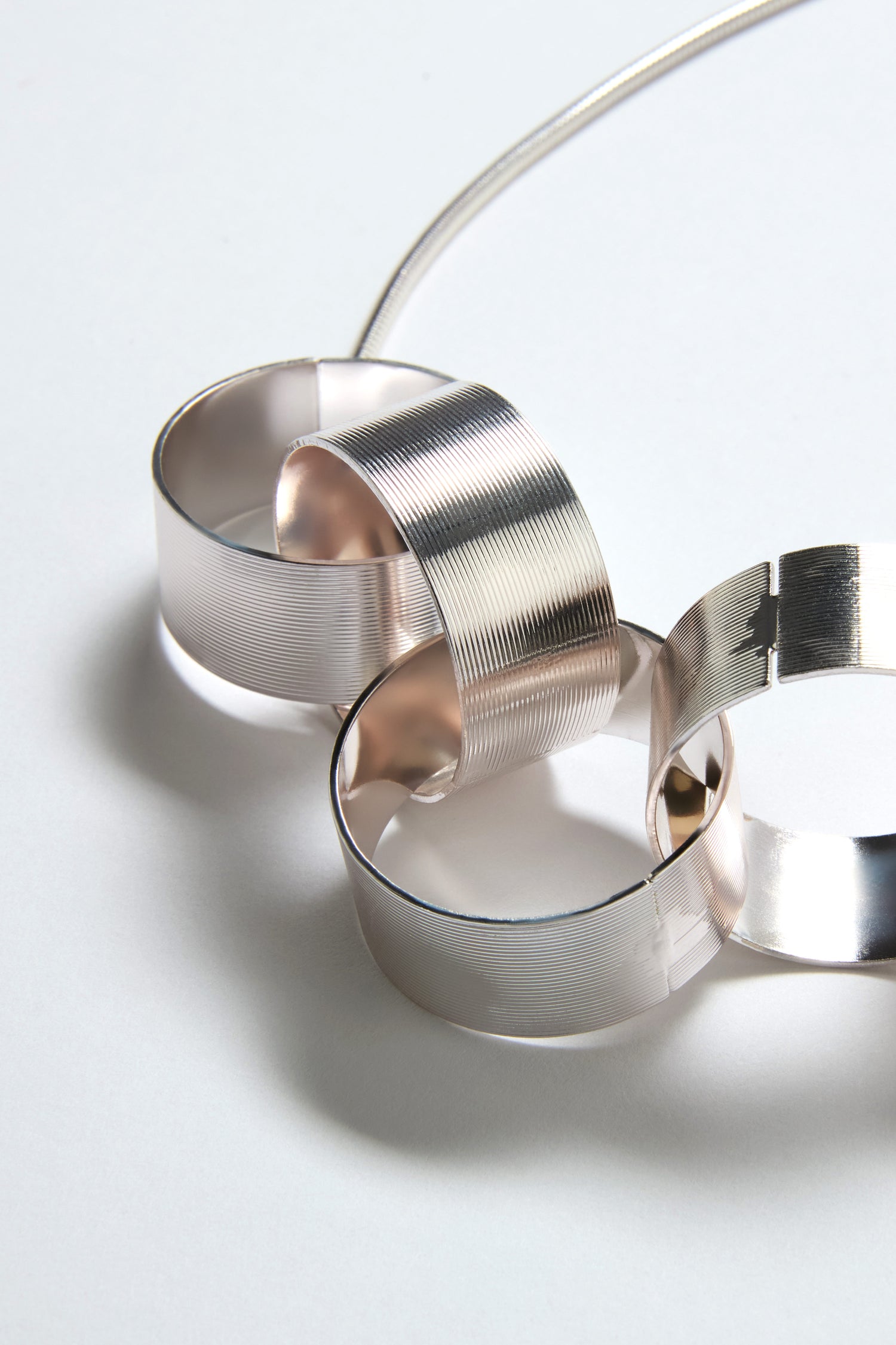 Three ribbed, brushed silver rings, interlocked and connected by a delicate chain, rest on a light surface—an elegant sculptural detail inspired by the Affinity Hoop Necklace.