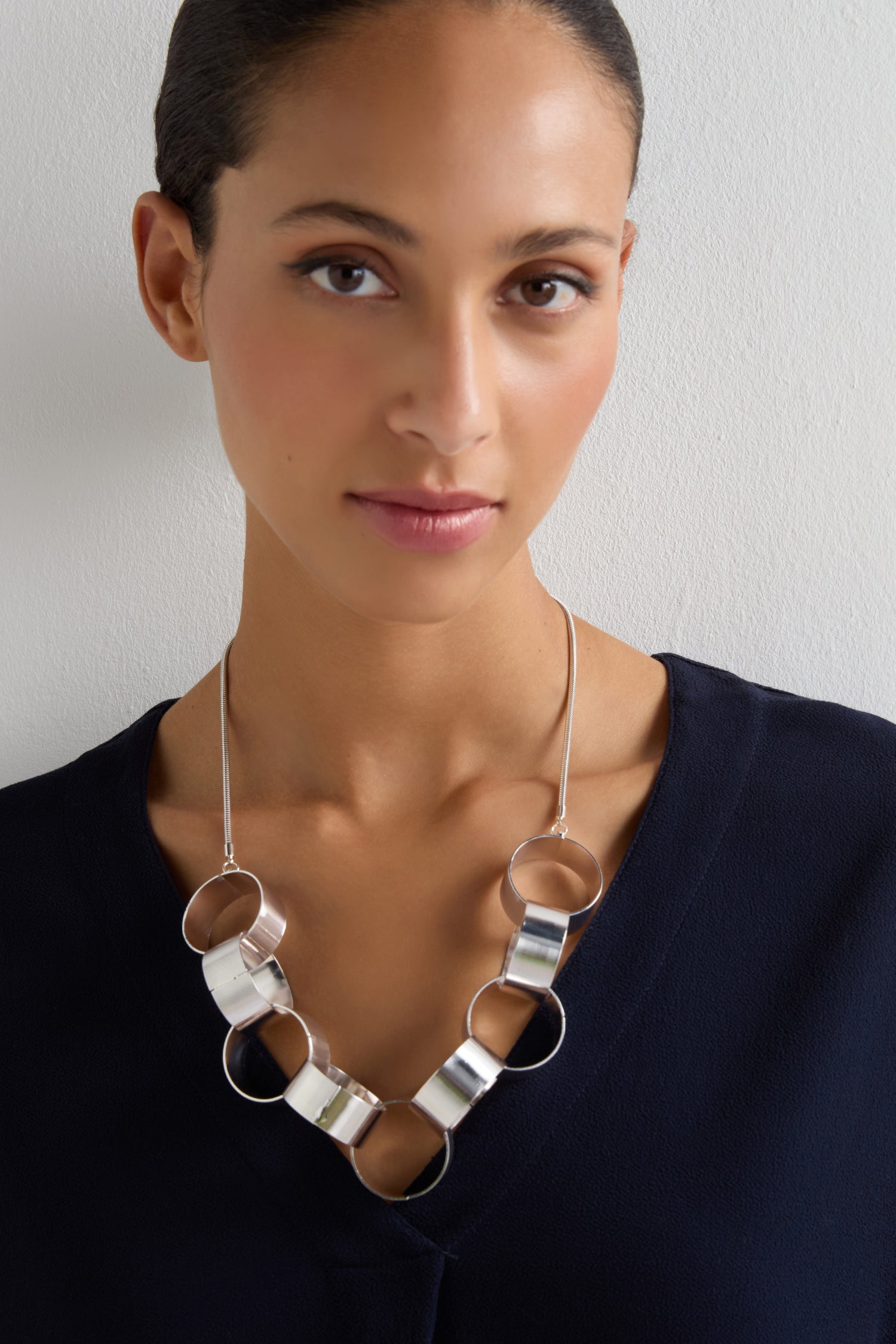 A woman wearing a navy top and the Affinity Hoop Necklace—featuring large interlocking circular links with a brushed silver finish—stands against a plain light background.