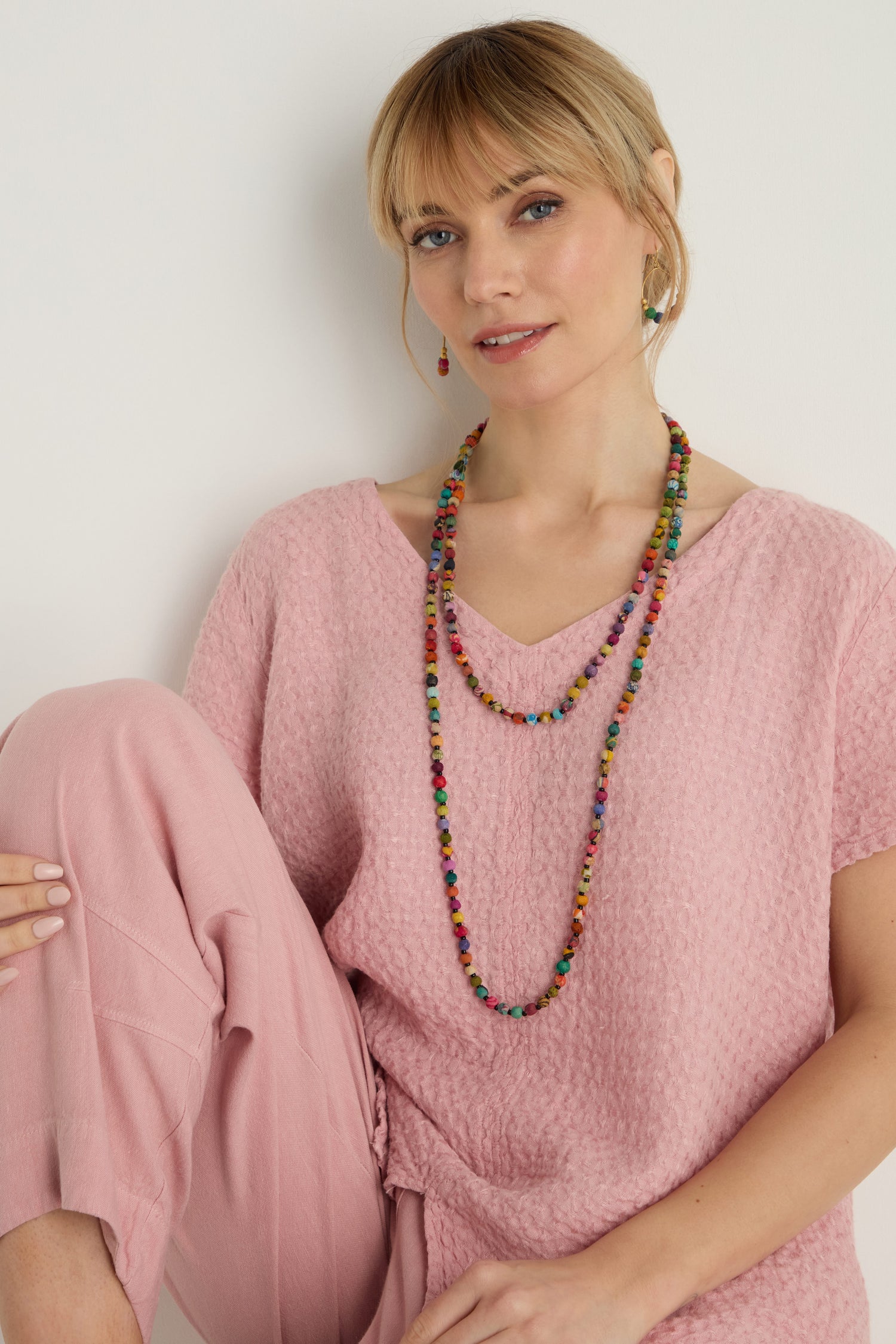 Woman wearing a textured pink top and pants, accessorized with the Kantha Long Bead Spheres Necklace and matching earrings, sitting against a plain white background.