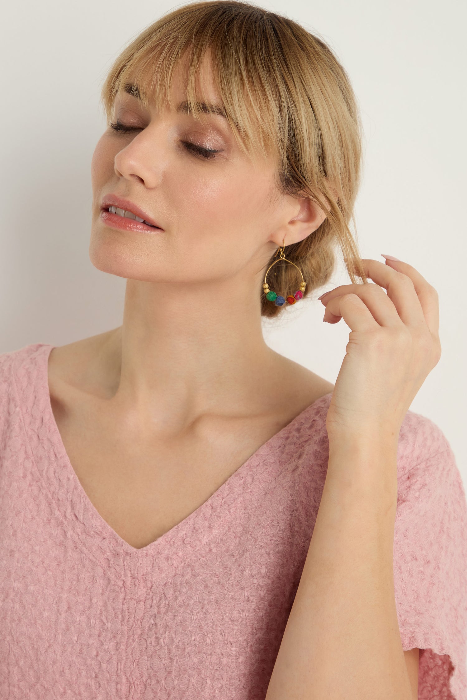 A woman in a textured pink top touches her hair, displaying the Kantha Gilded Hoop Earrings—handcrafted gold hoops embellished with vibrant beads—while posing against a plain light background.