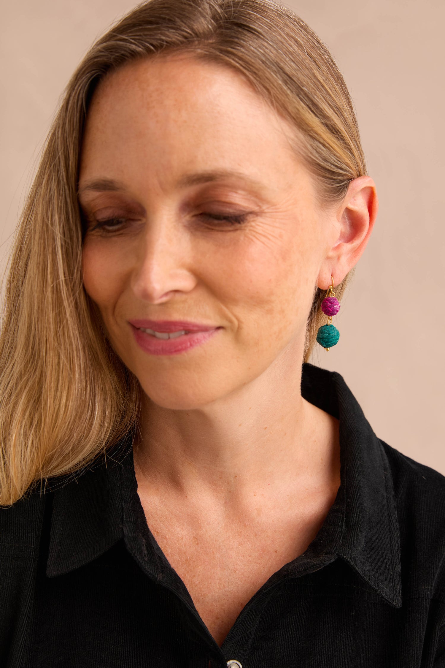 Woman with long blonde hair, wearing pink lipstick, a black collared shirt, and vibrant Kantha Double Spheres Earrings made of cotton-wrapped beads, looking down with a slight smile.