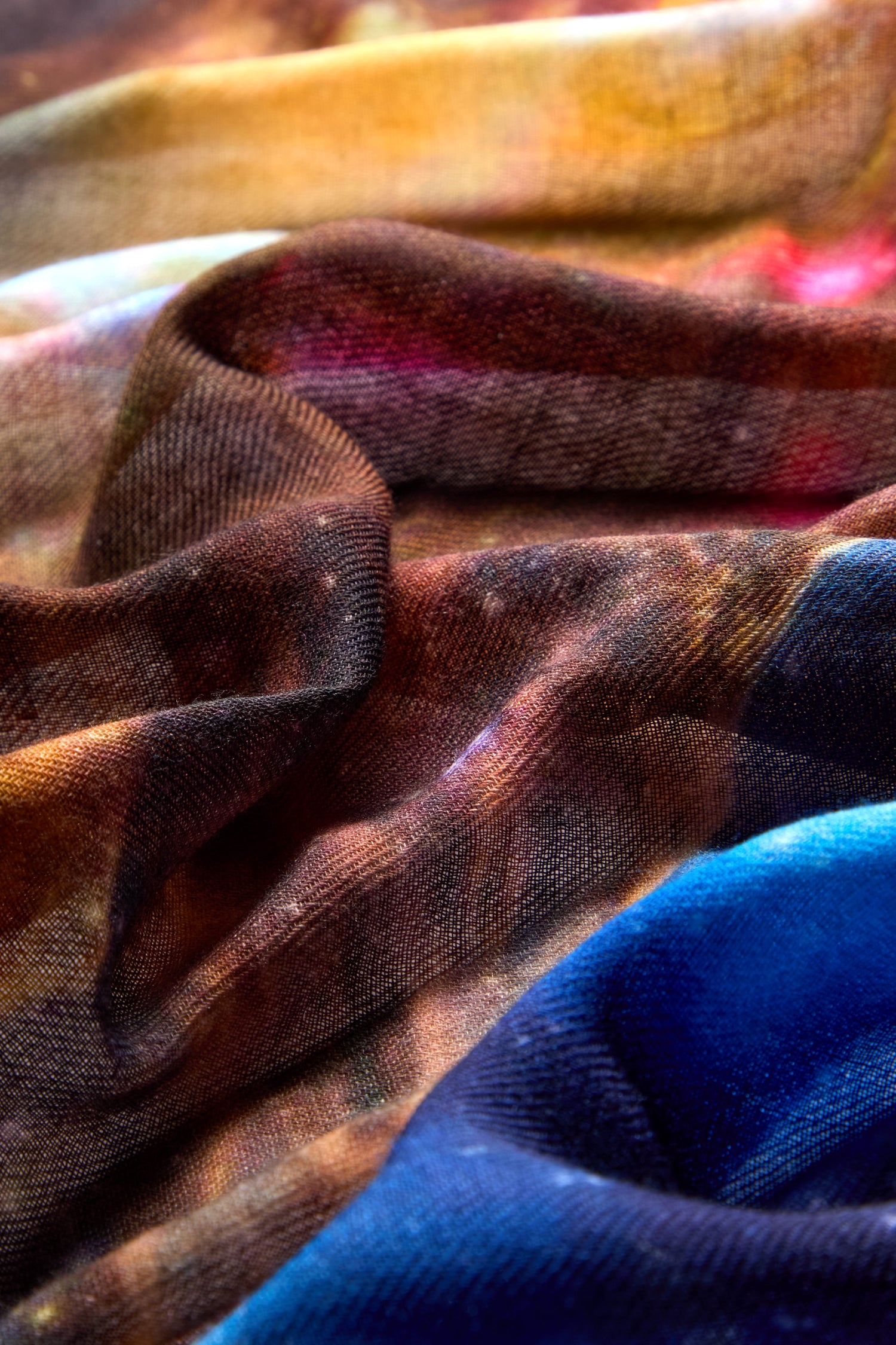 Close-up of a colorful, flowing fabric with a mix of brown, blue, and yellow hues. The texture appears soft and slightly transparent, embodying celestial elegance. This **Cosmic Sky Print Wool Scarf** exudes a captivating charm in every detail.