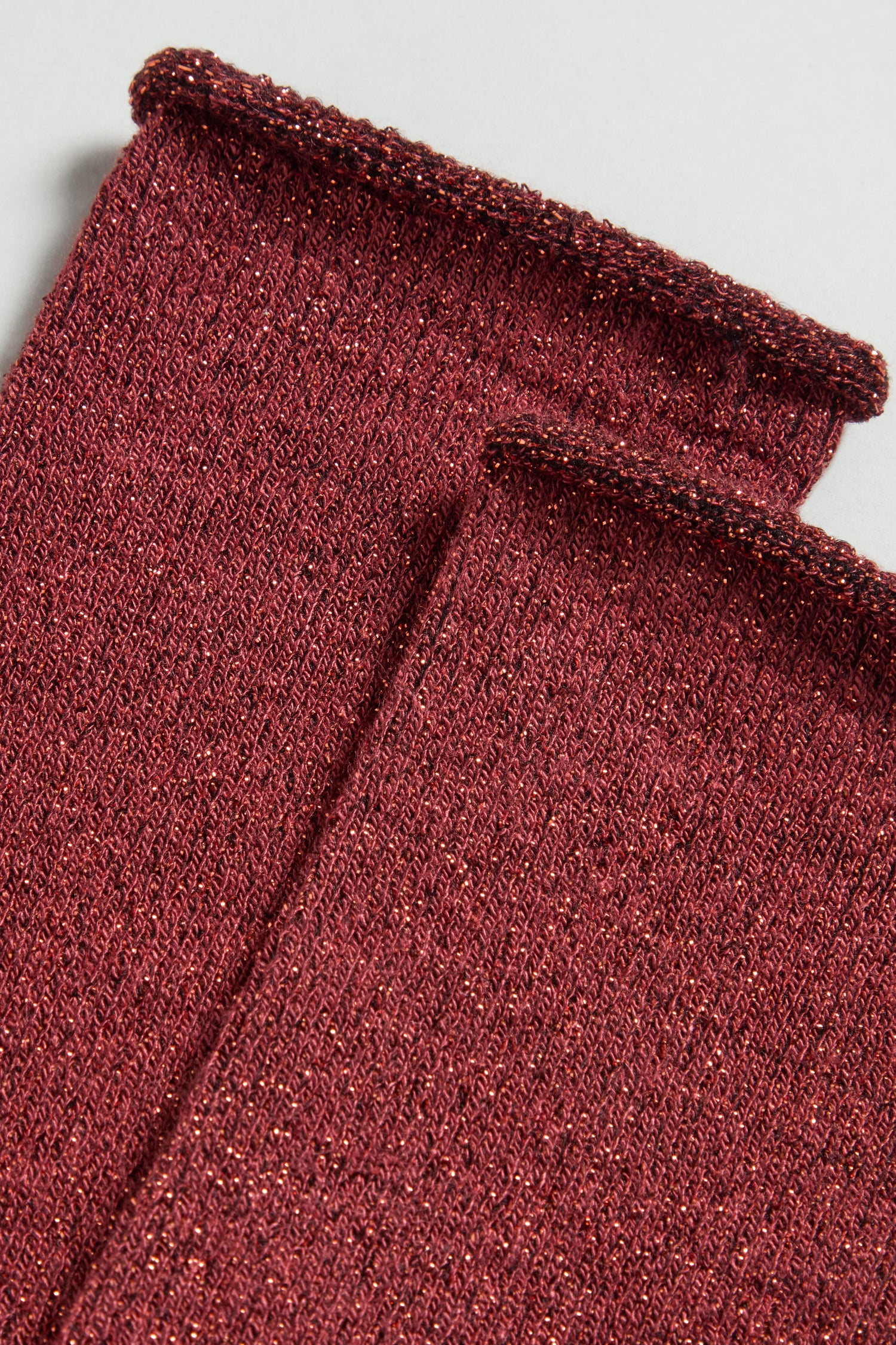 Close-up of two Bamboo Sparkle Red Socks in maroon knit with metallic threads, featuring rolled hems along the top, are laid flat on a light background.