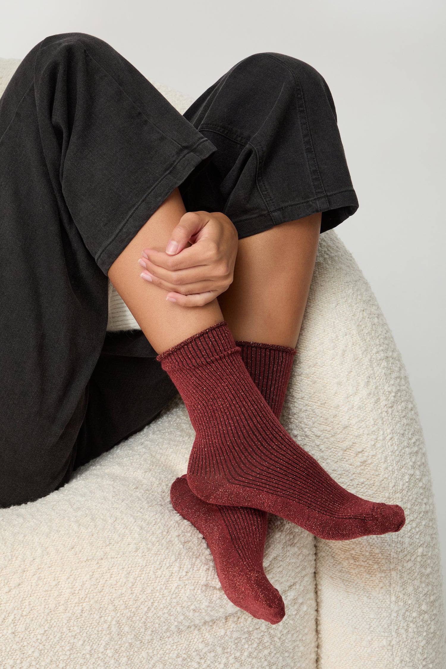 A person sits on a white textured chair with their legs crossed, wearing black pants and Bamboo Sparkle Red Socks.
