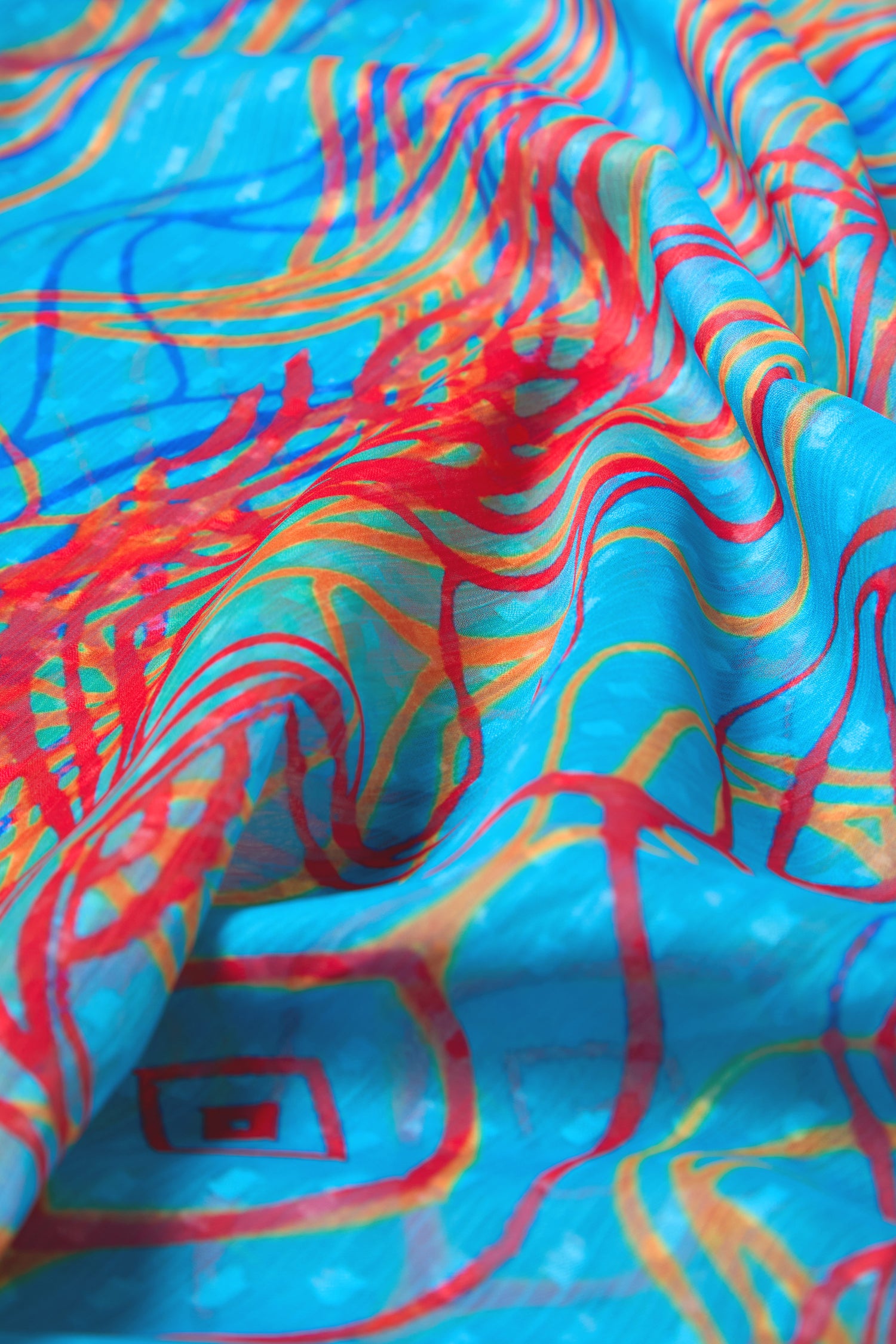 Close-up of the Twirl Print Dobby Scarf, featuring lightweight blue fabric with red, orange, and yellow abstract swirl patterns; folds highlight the scarf’s texture.