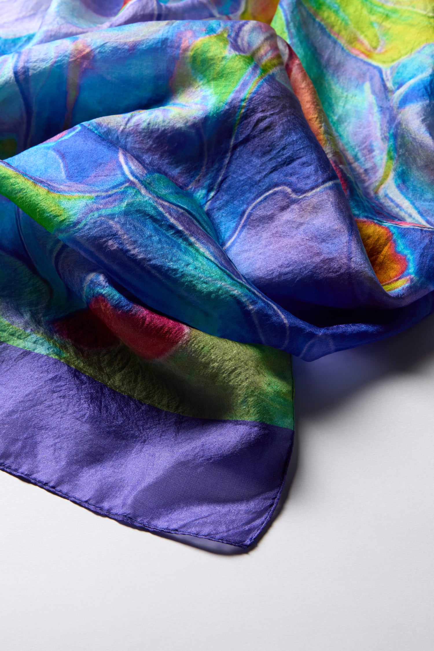 The Tiffany Lamp Silk Scarf, with its luxurious silk and vibrant shades of blue, green, red, and purple, elegantly crumples on a white surface.