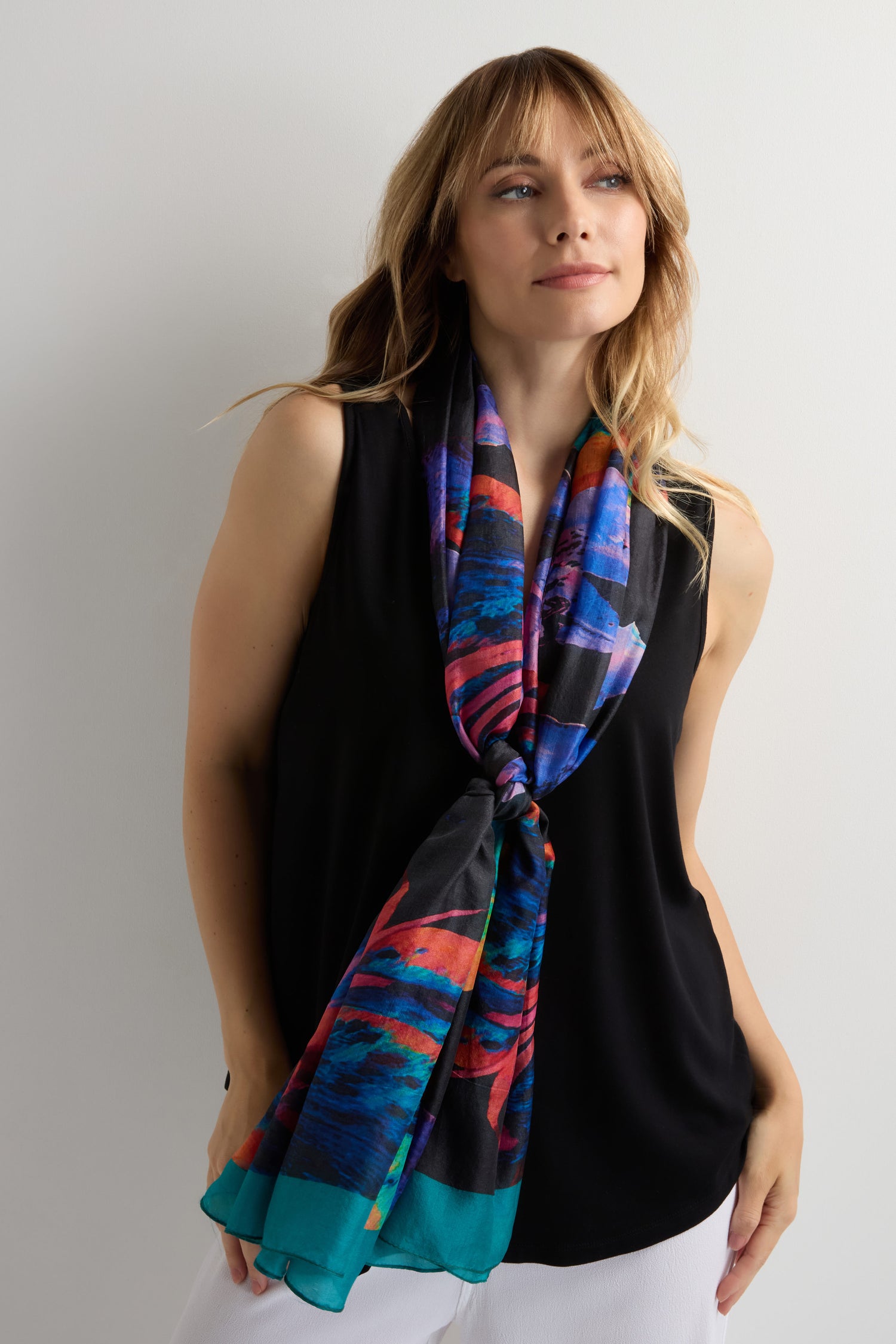 A woman in a sleeveless black top and white pants poses with the Orchid Silk Scarf, a colorful and modern elegant accessory, tied stylishly around her neck.