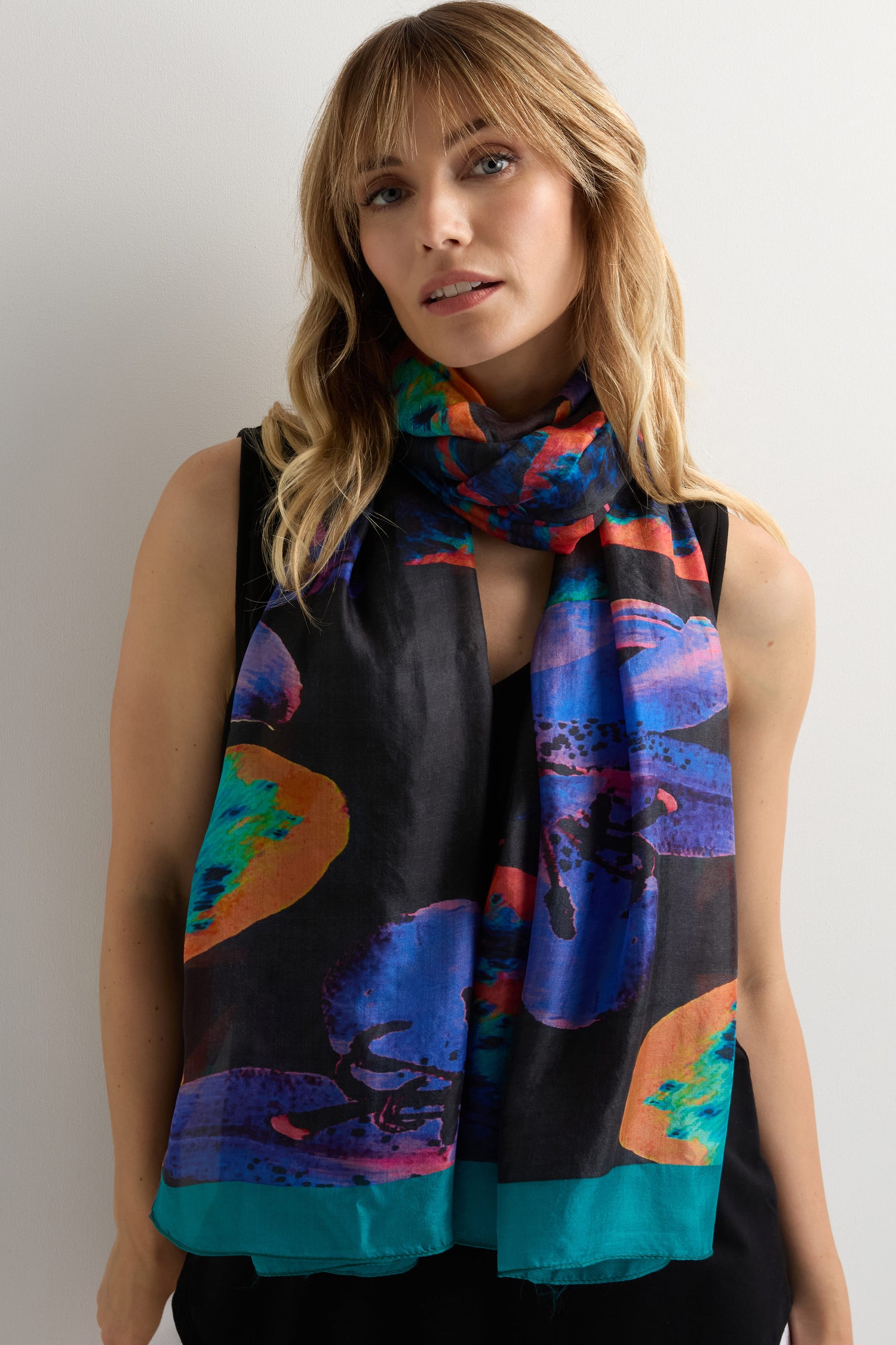 A woman wears a black sleeveless top and the Orchid Silk Scarf, featuring blue, orange, and green abstract patterns, standing against a plain light background.
