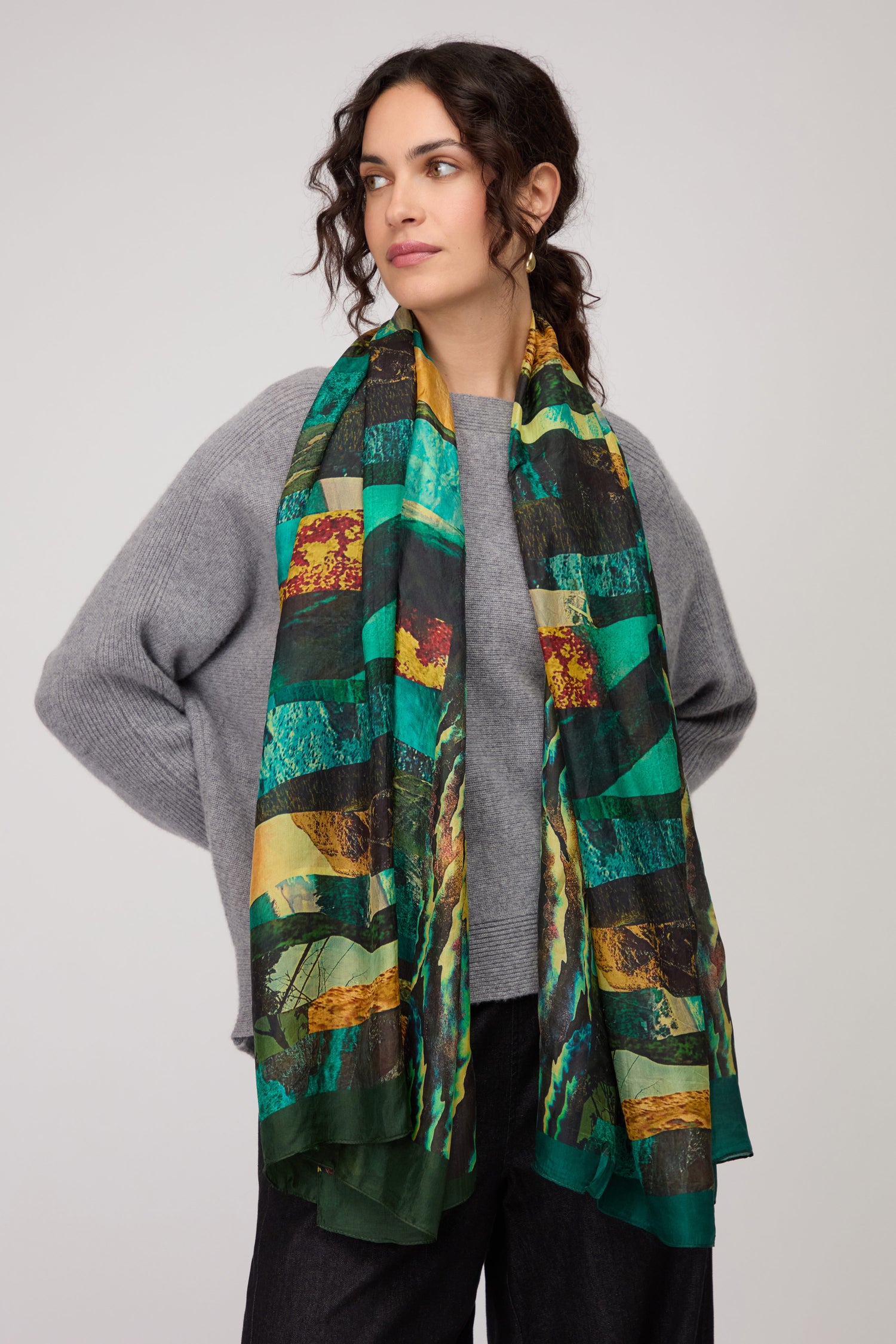 A woman wears a grey sweater and black pants, elegantly draping the Northern Lights Silk Scarf—long, multicolored in green and yellow with an abstract design—over her shoulders.
