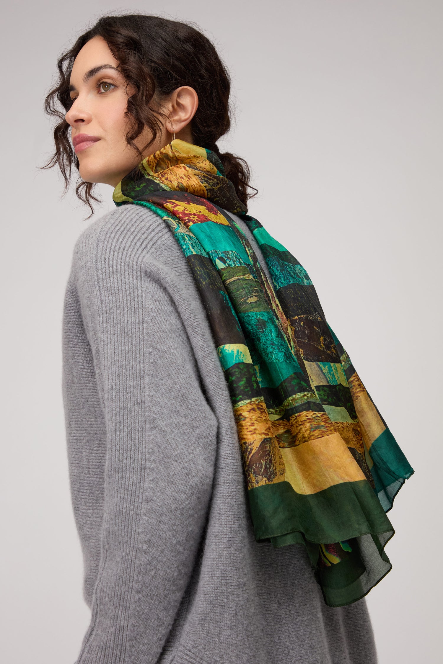 A woman in a gray sweater wears the Northern Lights Silk Scarf, featuring an abstract multicolored design, draped over her shoulder as she looks sideways.