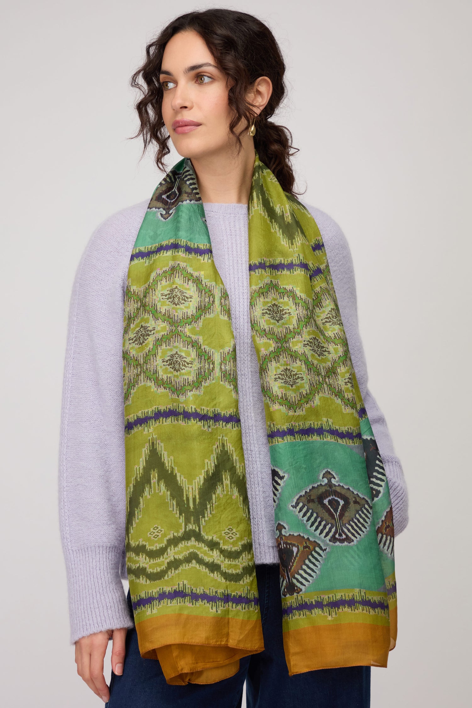 A woman stands against a plain background, wearing a light purple sweater, blue jeans, and the vibrant Aztec Silk Scarf draped around her neck.