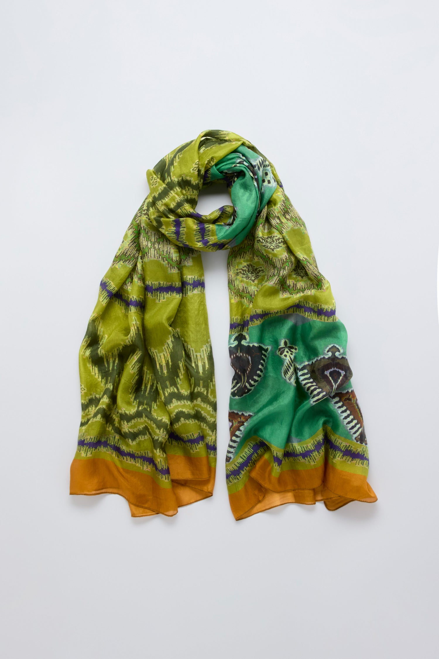 The Aztec Silk Scarf features tribal-inspired abstract designs and animal-like figures in green and yellow with brown edges, shown laid flat on a white background.