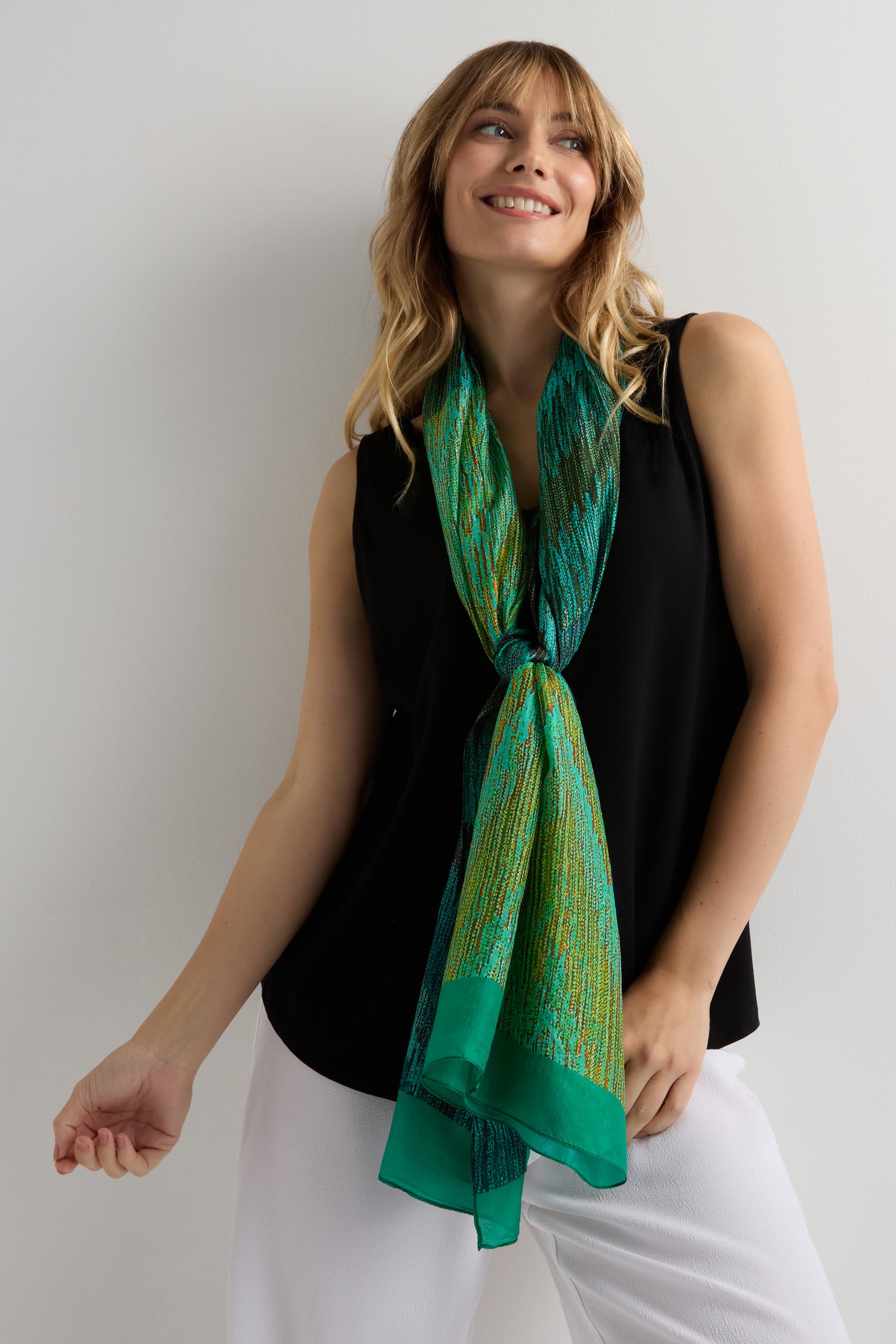 Smiling woman stands against a light wall, wearing a sleeveless black top, white pants, and the Shaded Sketch Green and Blue Silk Scarf with an abstract print.