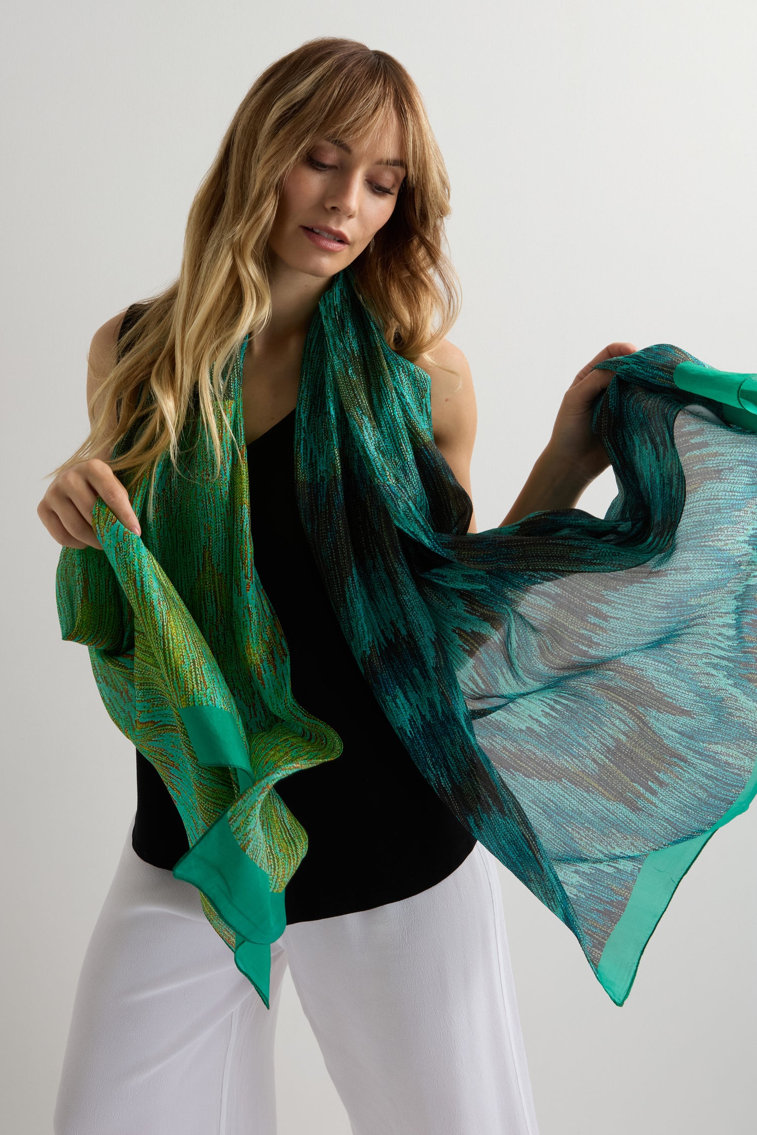 A woman with long blonde hair, dressed in a sleeveless black top and white pants, holds the Shaded Sketch Green and Blue Silk Scarf with an abstract print against a plain light background.