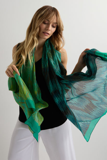 A woman with long blonde hair, dressed in a sleeveless black top and white pants, holds the Shaded Sketch Green and Blue Silk Scarf with an abstract print against a plain light background.