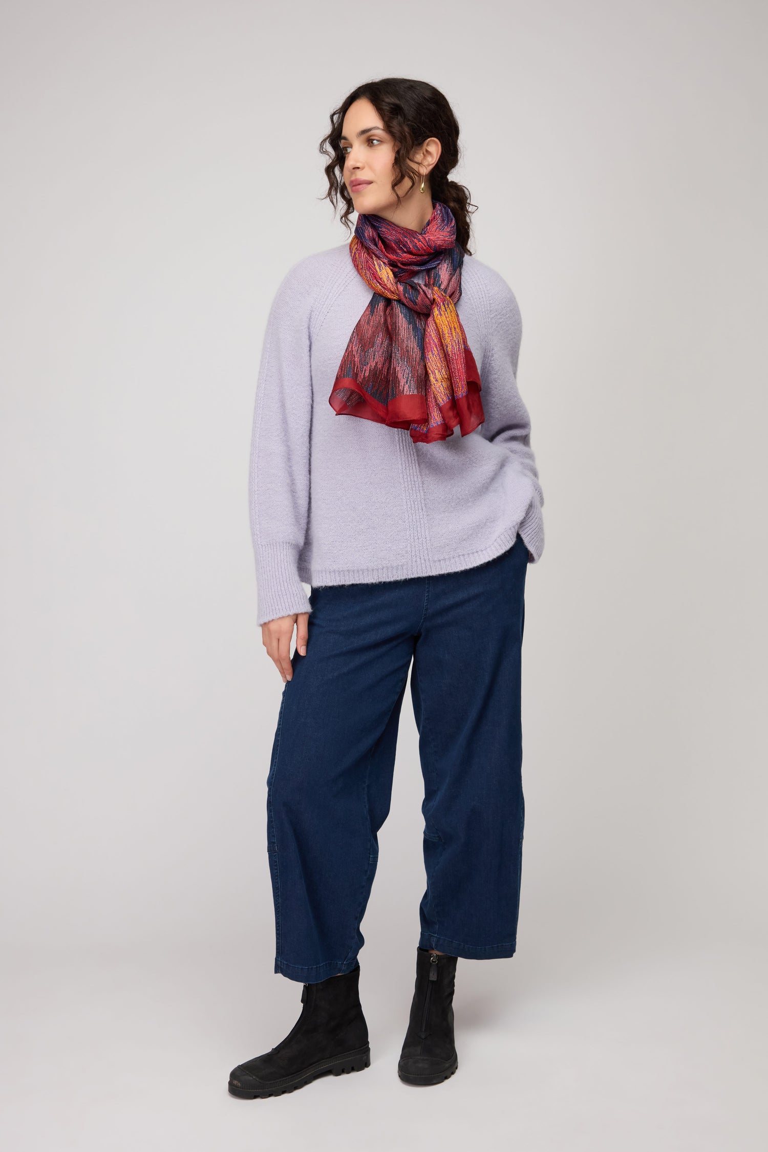 Woman standing against a plain background, wearing a light sweater, dark wide-leg pants, black boots, and the Shaded Sketch Silk Scarf—an eye-catching lightweight accessory—with one hand in her pocket.