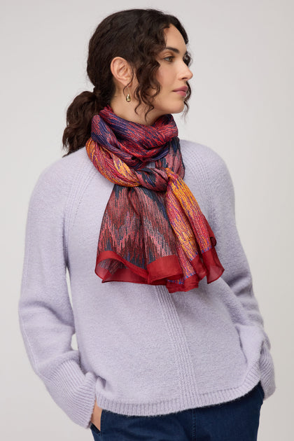 A woman with curly dark hair wears a light purple sweater, blue jeans, and the Shaded Sketch Silk Scarf, standing against a plain light background.