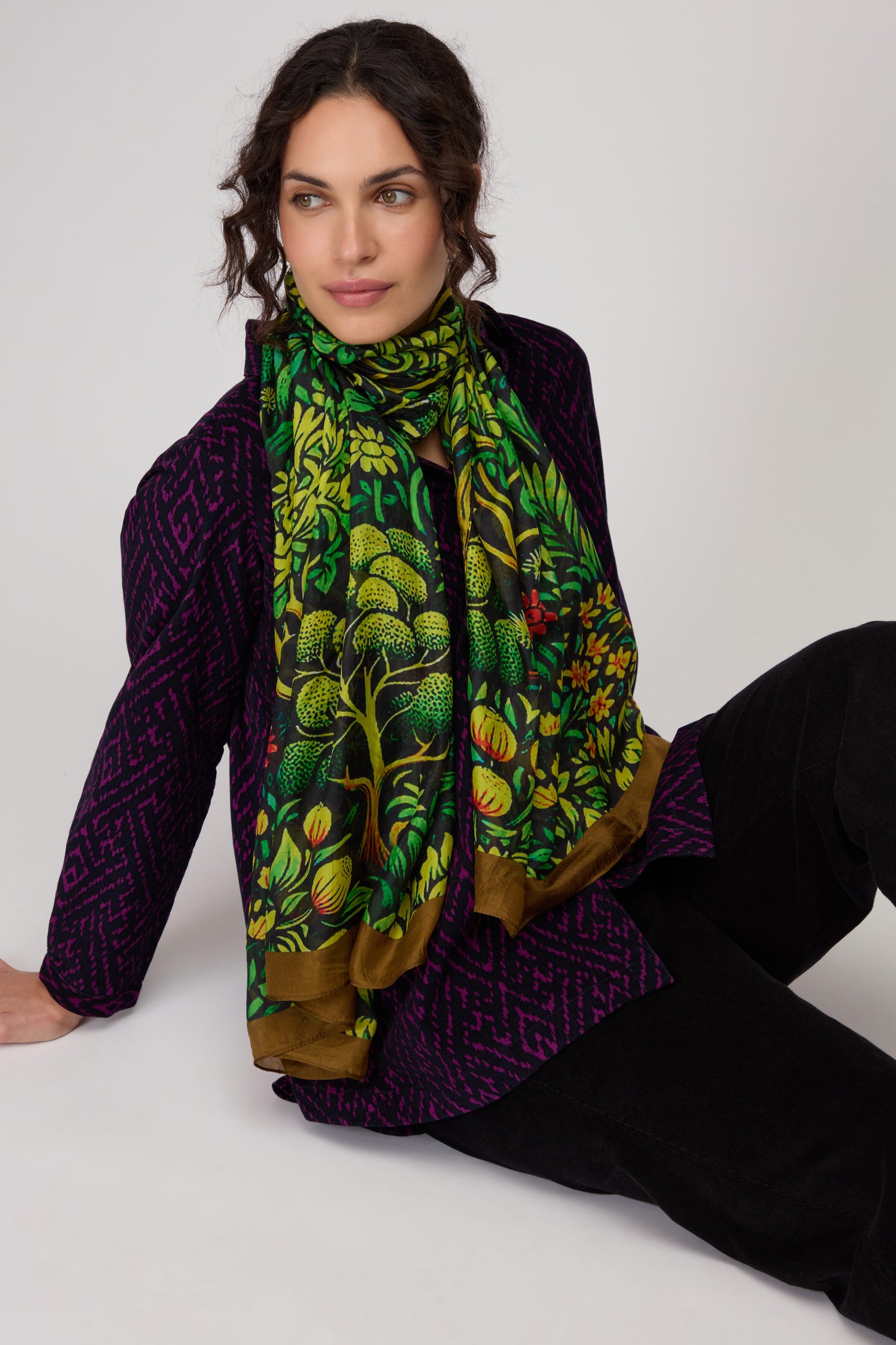 A woman with wavy dark hair wears a dark patterned jacket and the Autumnal Garden Silk Scarf, its vibrant floral print echoing autumn foliage, as she sits on the floor against a plain light background.