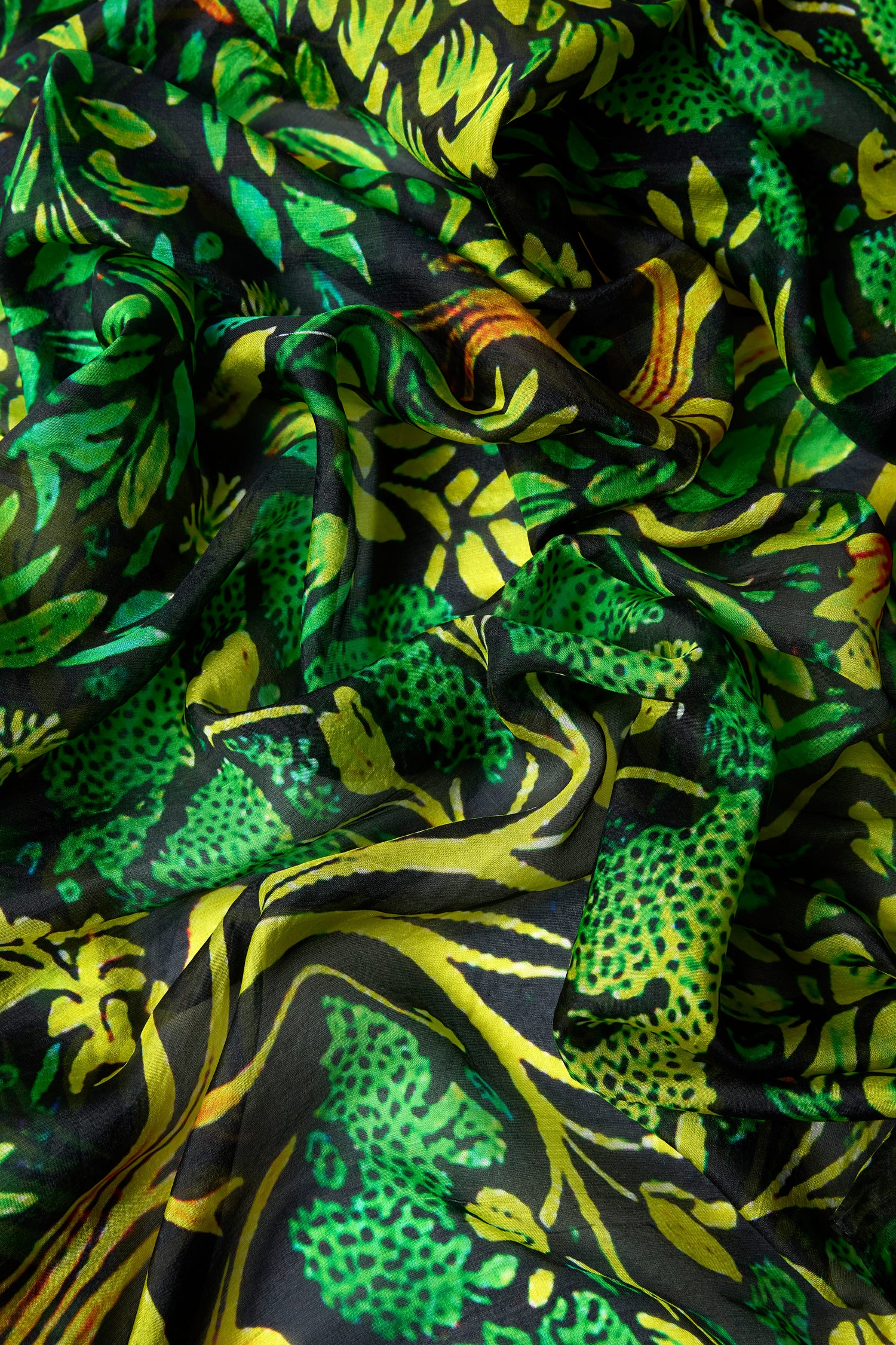 A close-up of luxurious crumpled fabric showcases the Autumnal Garden Silk Scarf, featuring a vibrant green, yellow, and black floral design reminiscent of autumn foliage.