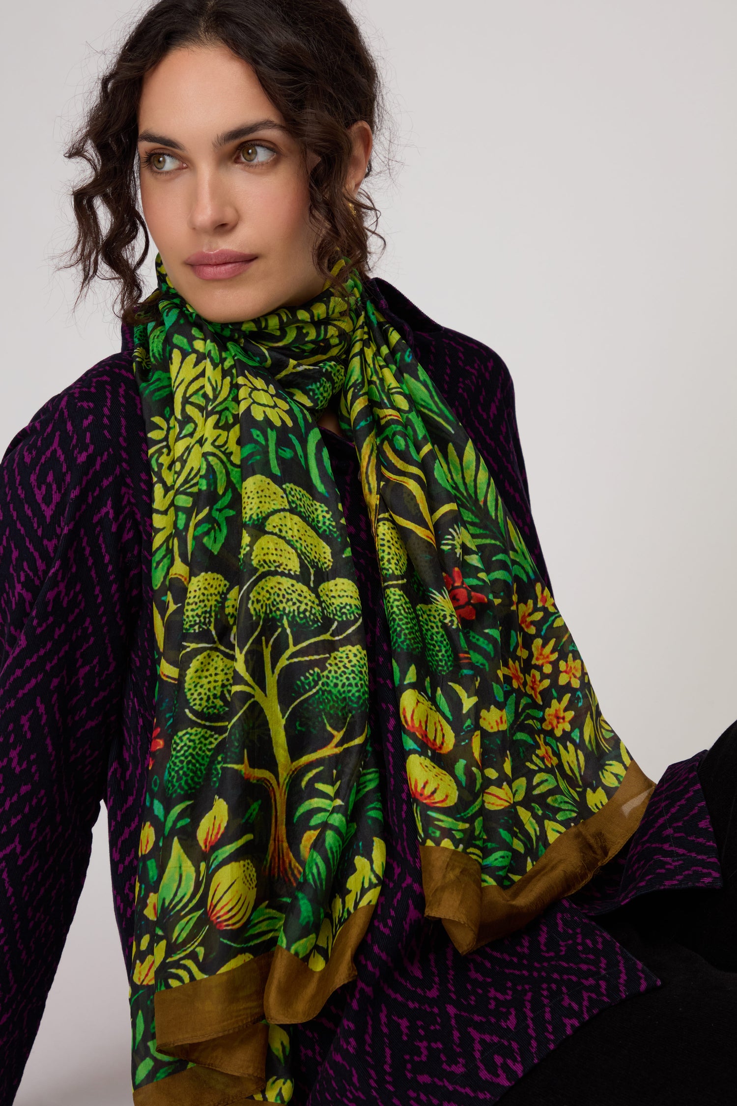 Woman with curly hair wearing a purple patterned top and the Autumnal Garden Silk Scarf, featuring a botanical print, sitting against a plain background.