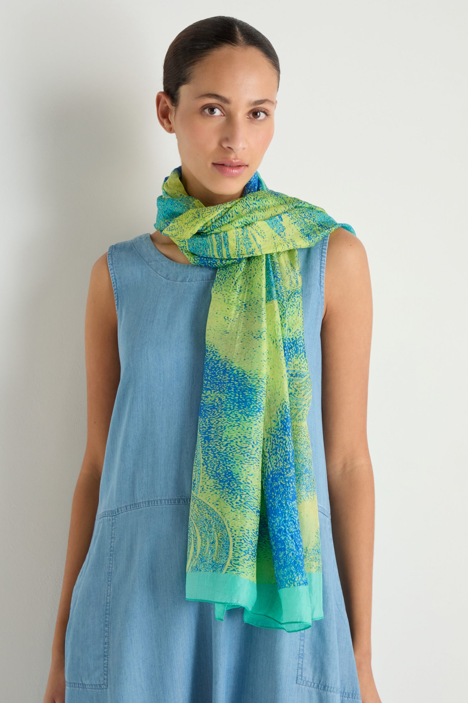A woman in a sleeveless light blue dress stands against a plain light background, styled with the Sky Dance Silk Scarf—a lightweight accessory featuring green and blue patterns.