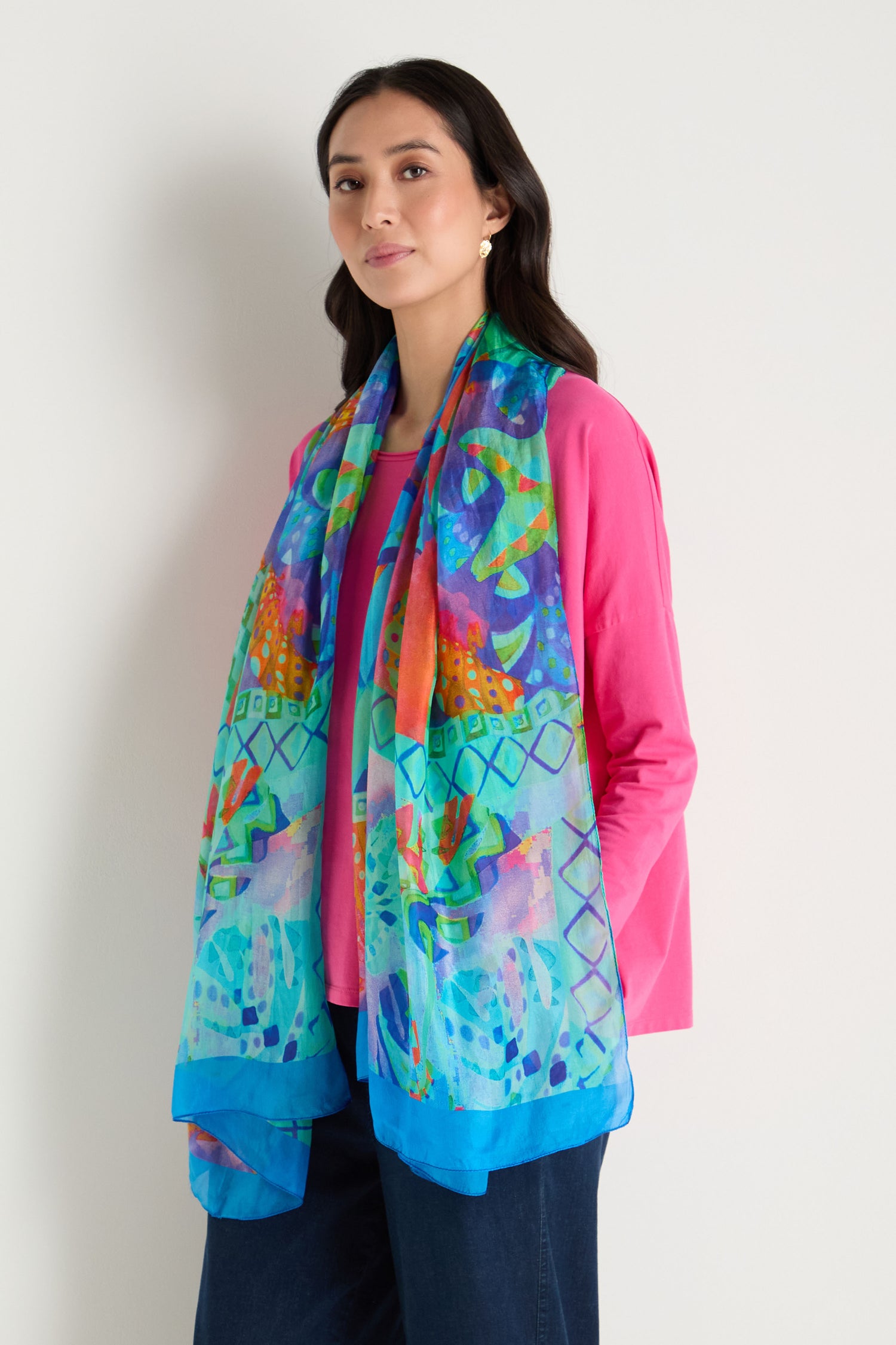 A woman stands against a plain light background, wearing a bright pink long-sleeve top, dark pants, and the Ocean Garden Silk Scarf featuring blue, green, and orange tones in a lively coastal print.